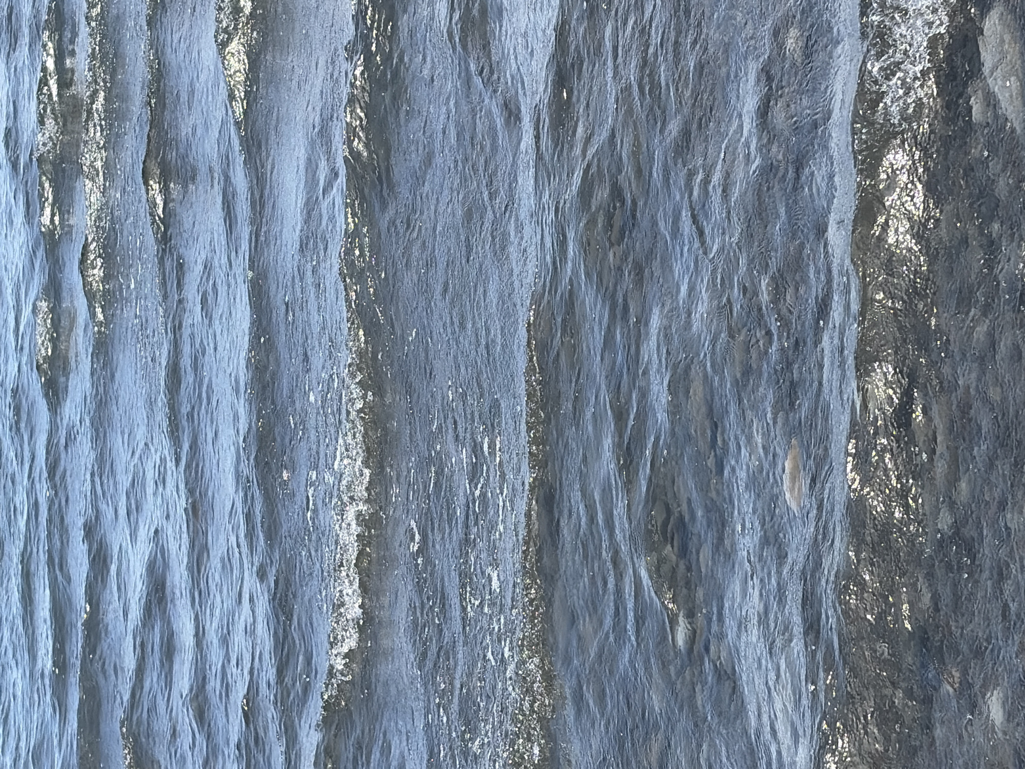 The image shows a close-up view of choppy water, likely a lake or ocean, with small waves rippling across the surface. The water is a deep blue-gray, and sunlight glints off the crests of the waves, creating sparkling highlights. The bottom of the body of water is visible in places, appearing to be covered with small stones and pebbles. There is one light-colored, round object in the water, possibly a buoy or a floating piece of debris. The scene is natural and tranquil, with no discernible man-made structures or human activity. The location is Unknown, Unknown.