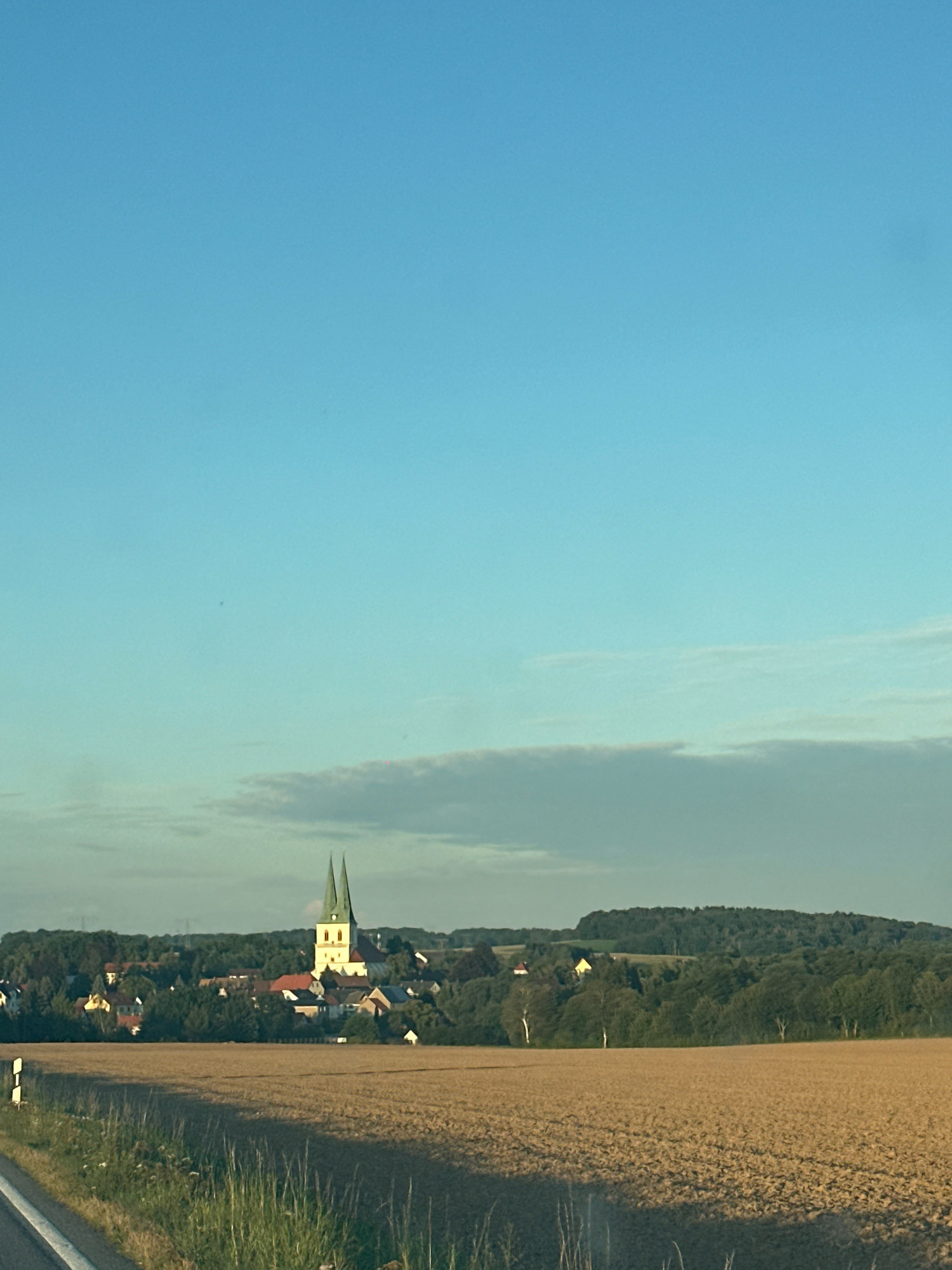 The image depicts a rural landscape under a clear blue sky. In the middle ground, a village is nestled amongst trees, with a prominent church featuring two green spires at its center. The foreground shows a tilled field, likely farmland, with a dirt road or path running along its left edge. The scene appears to be taken from a slightly elevated position, possibly from a vehicle, looking towards the village and beyond. The lighting suggests it is either early morning or late afternoon, given the warm tone of the field and the soft shadows. The location is identified as Göda - Hodźij, Germany.