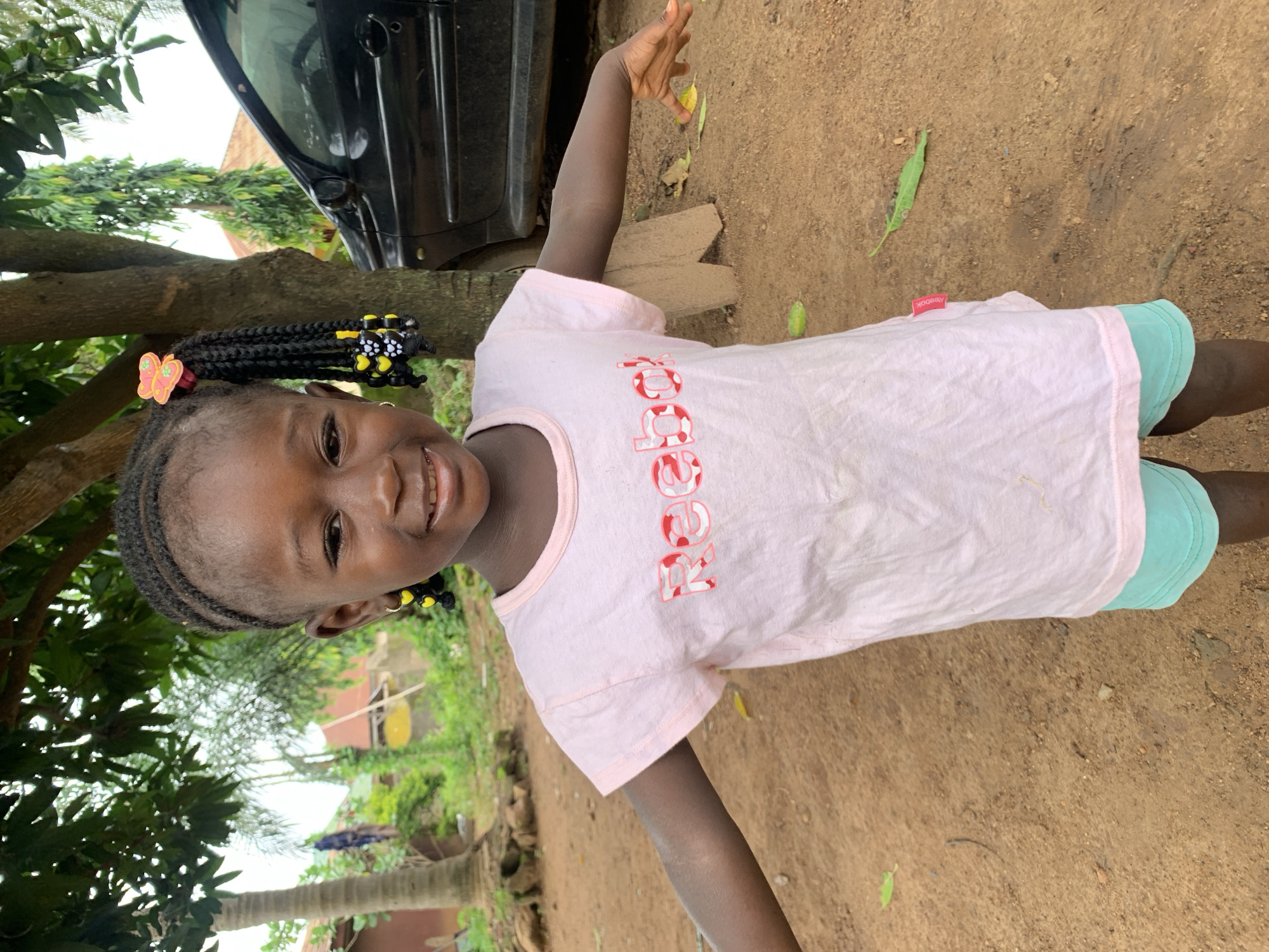 A young Black girl with braided hair adorned with yellow heart-shaped beads and a pink butterfly clip stands in a dirt yard, facing forward and smiling. She wears a pale pink t-shirt with a red and white logo that appears to say "Reebok" and light blue shorts. Her arms are outstretched to her sides. Behind her, a black car is parked. The background features trees, foliage, and a building, suggesting an outdoor, possibly rural or suburban setting. The ground is covered in reddish-brown dirt.