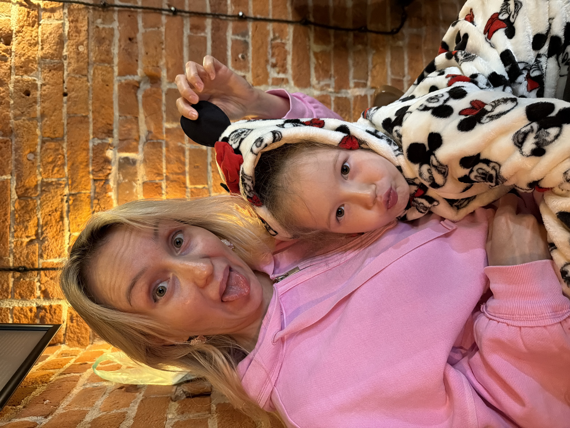 A woman and a child are posing for a selfie, against the backdrop of a brick wall. The woman, with blonde hair, is wearing a pink hoodie and sticking her tongue out with wide eyes. The child, wearing a hooded onesie with a Mickey Mouse pattern, is positioned on top of the woman. The hood of the onesie has Mickey Mouse ears attached. The location is a restaurant in Moscow, Russia.