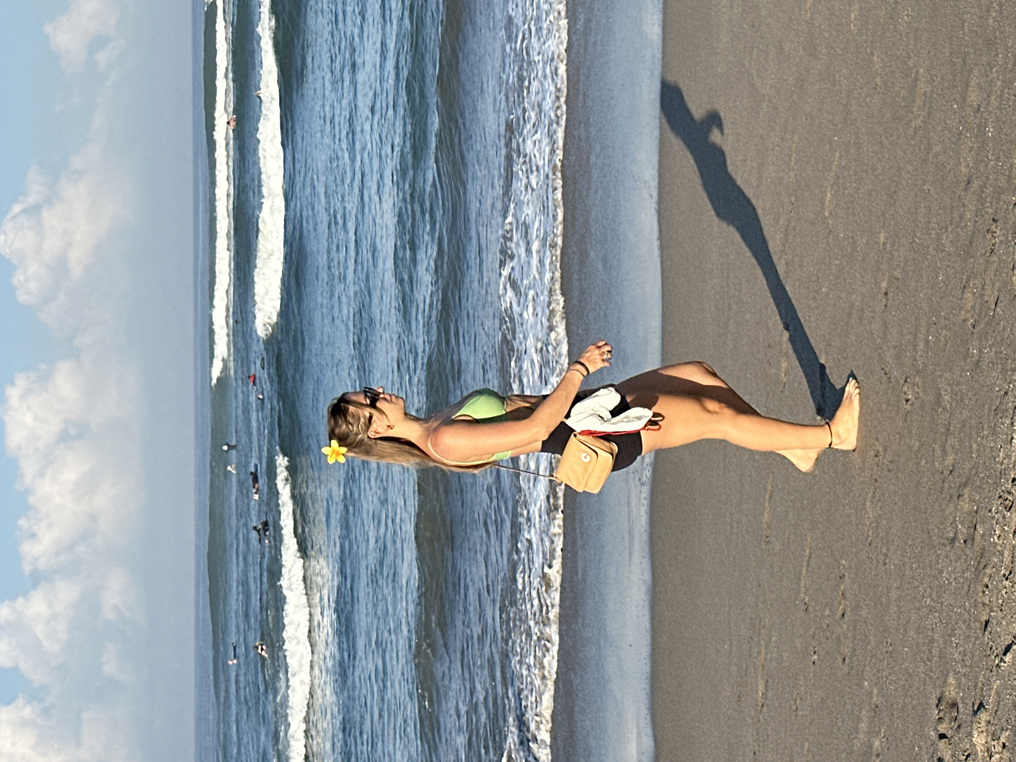 A woman walks along the beach in North Kuta, Indonesia, with the ocean and waves behind her. She has a yellow flower in her hair, sunglasses, and is wearing a green bikini top and black shorts. A tan crossbody bag is slung over her shoulder, and she is carrying a white garment. The sand is dark and wet, with some footprints visible. The sun is casting a long shadow of the woman on the sand. In the background, several surfers can be seen in the water, waiting for waves. The sky is blue with scattered white clouds.
