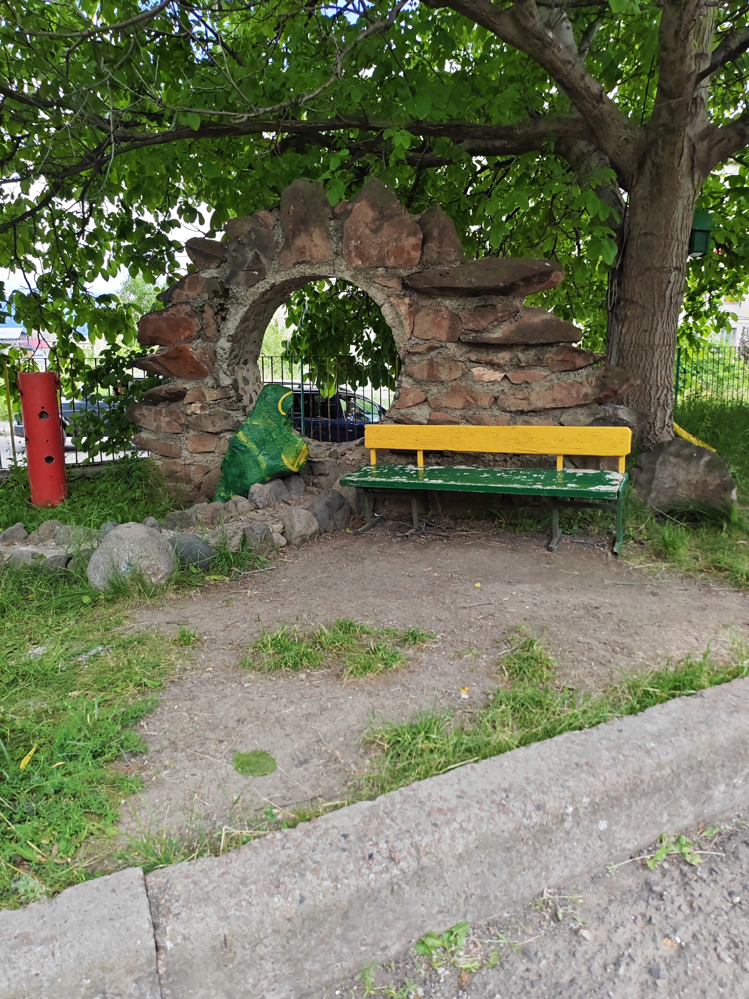 In Vanadzor, Armenia, an outdoor scene features a decorative stone archway and a bench. The archway is constructed with rough, reddish-brown stones and has an opening through which a glimpse of a dark-colored car is visible. To the left of the arch, a green, cartoonish figure resembling a dinosaur or dragon is seated among rocks. In front of the archway, a green bench with a yellow backrest sits on a dirt and grass area. A large tree with green foliage provides shade over the entire scene. A red cylindrical object is also present on the far left. The ground in the foreground is a mix of dirt, gravel, and grass, with a concrete curb running along the bottom edge of the image. A metal fence is visible behind the archway and bench.