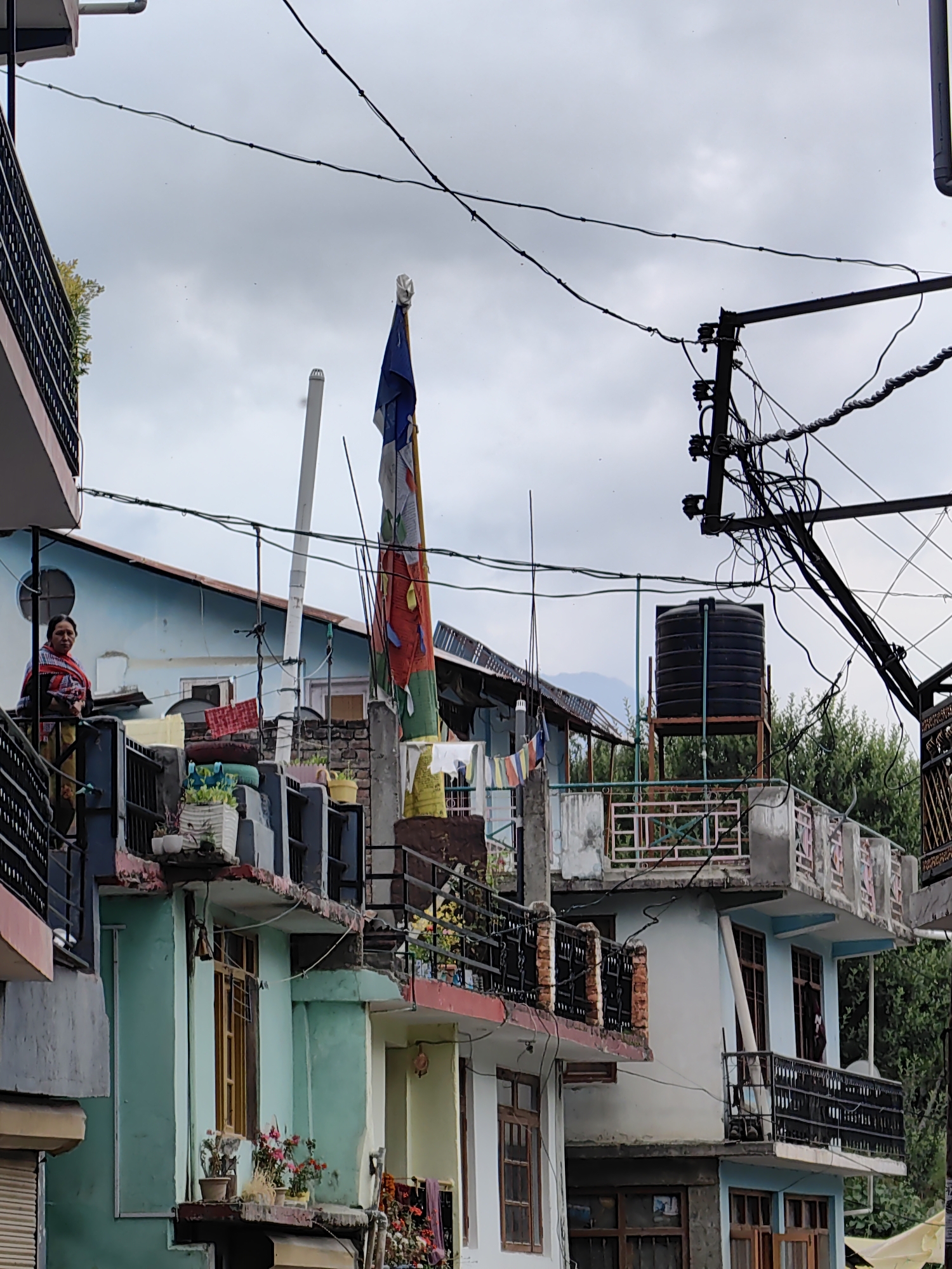 The image depicts a residential area in Vashisht, India, with a focus on multi-story buildings and the surrounding infrastructure. The sky is overcast with a cloudy, grey hue.
In the foreground, to the left, a woman wearing a red and black patterned shawl over a yellow garment stands on a balcony, looking slightly off-camera. Her arms are clasped in front of her. The balcony she is on is part of a building with a dark railing and a pink trim.
Dominating the center of the image is a tall, narrow flag pole with a flag partially unfurled. The flag is a collection of colorful rectangular panels, predominantly white, blue, red, and green, attached together vertically. The flag appears to be a prayer flag, commonly seen in Buddhist regions. Beside the flag pole, a white pipe extends upwards.
Behind the flag, on a higher level, is a black water tank mounted on a metal stand. A complex network of black electrical wires, supported by poles and insulators, crisscrosses the upper portion of the frame, indicating the presence of electricity infrastructure.
The buildings in the scene exhibit varied architectural styles and color schemes. Some are painted in light blue and white, while others are a vibrant teal green. Many have balconies with dark railings. Windows with wooden frames are visible on several levels.
Potted plants and flowers are placed on some of the balconies and ledges, adding touches of greenery and color to the urban environment. In the mid-ground, laundry is hung out to dry on a clothesline between buildings.
The overall impression is that of a densely populated urban setting with a blend of traditional and functional elements, characteristic of a town like Vashisht, known for its spiritual sites and mountainous backdrop, although mountains are only faintly visible in the distance on this particular day. The scene is static, capturing a moment in time without significant action or interaction.