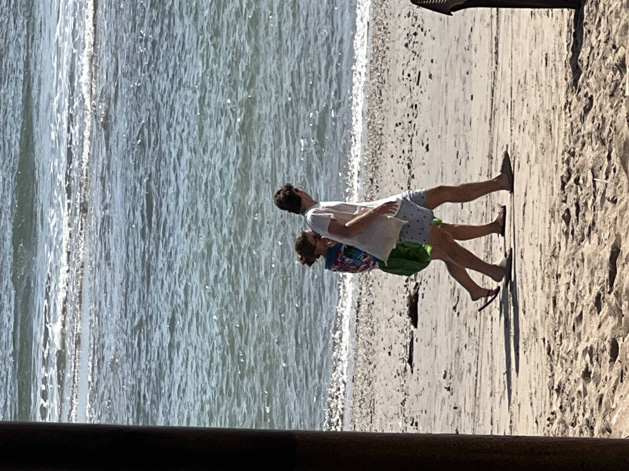 Two individuals walk along the shoreline in Kuta, Indonesia. A person in the foreground, facing right and partially obscured by a dark vertical pole on the left, wears a light-colored t-shirt and patterned shorts. To their left, a person walks towards the right, wearing a colorful garment over a green skirt and flip-flops. They are walking on a sandy beach with a scattering of shells and debris. The ocean, with gentle waves and white foam, stretches out behind them. The scene is viewed from a sheltered area, indicated by the dark pole.