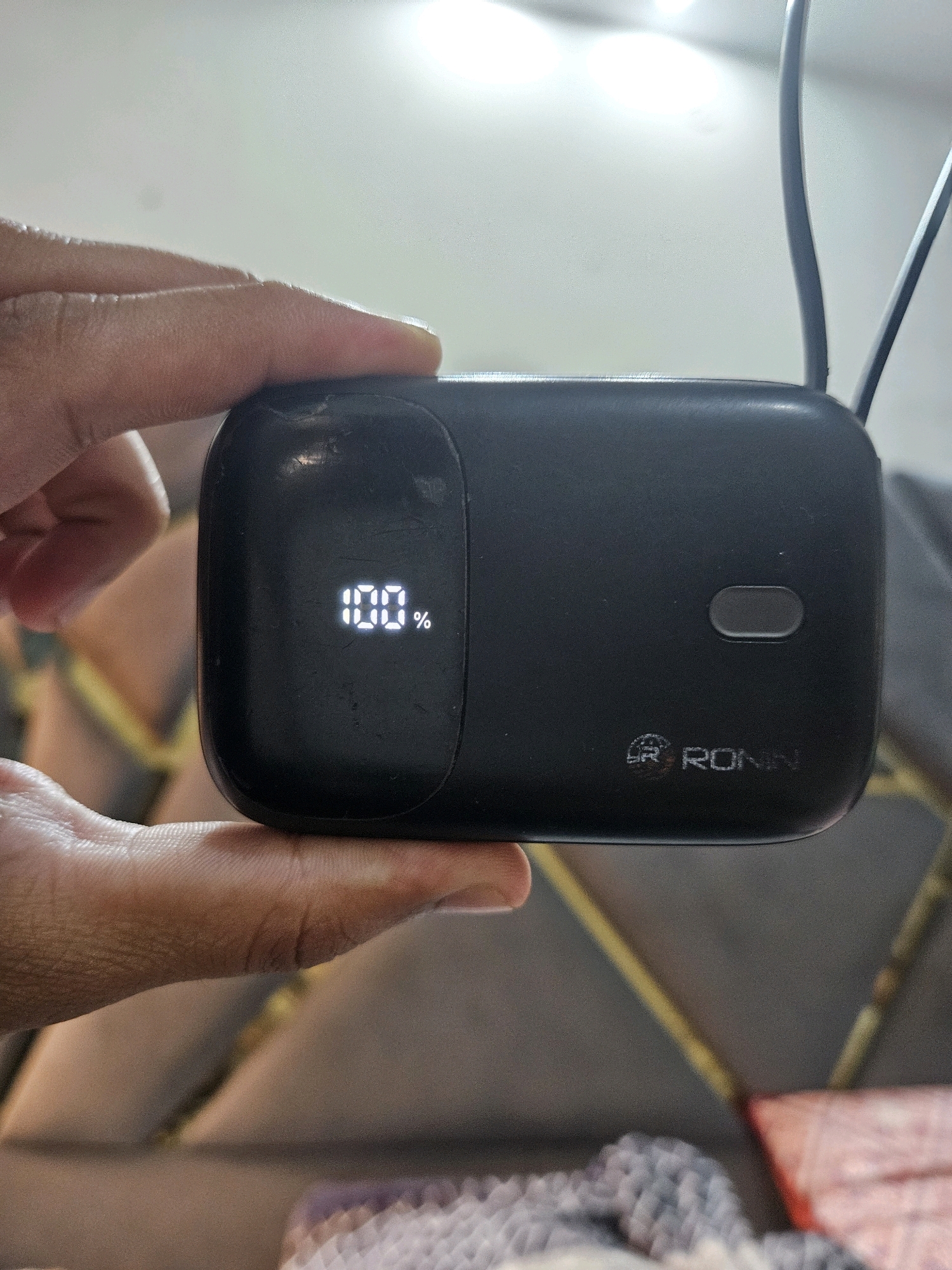 A close-up, top-down shot shows a black Ronin brand power bank held in a person's left hand. The power bank has a digital display showing "100%". A small, oval button is visible on the right side of the power bank. The background is blurred, suggesting it is a domestic environment, possibly a bedroom or living room, with a patterned upholstered surface beneath. The image is not taken in Jawad Avenue, Pakistan.