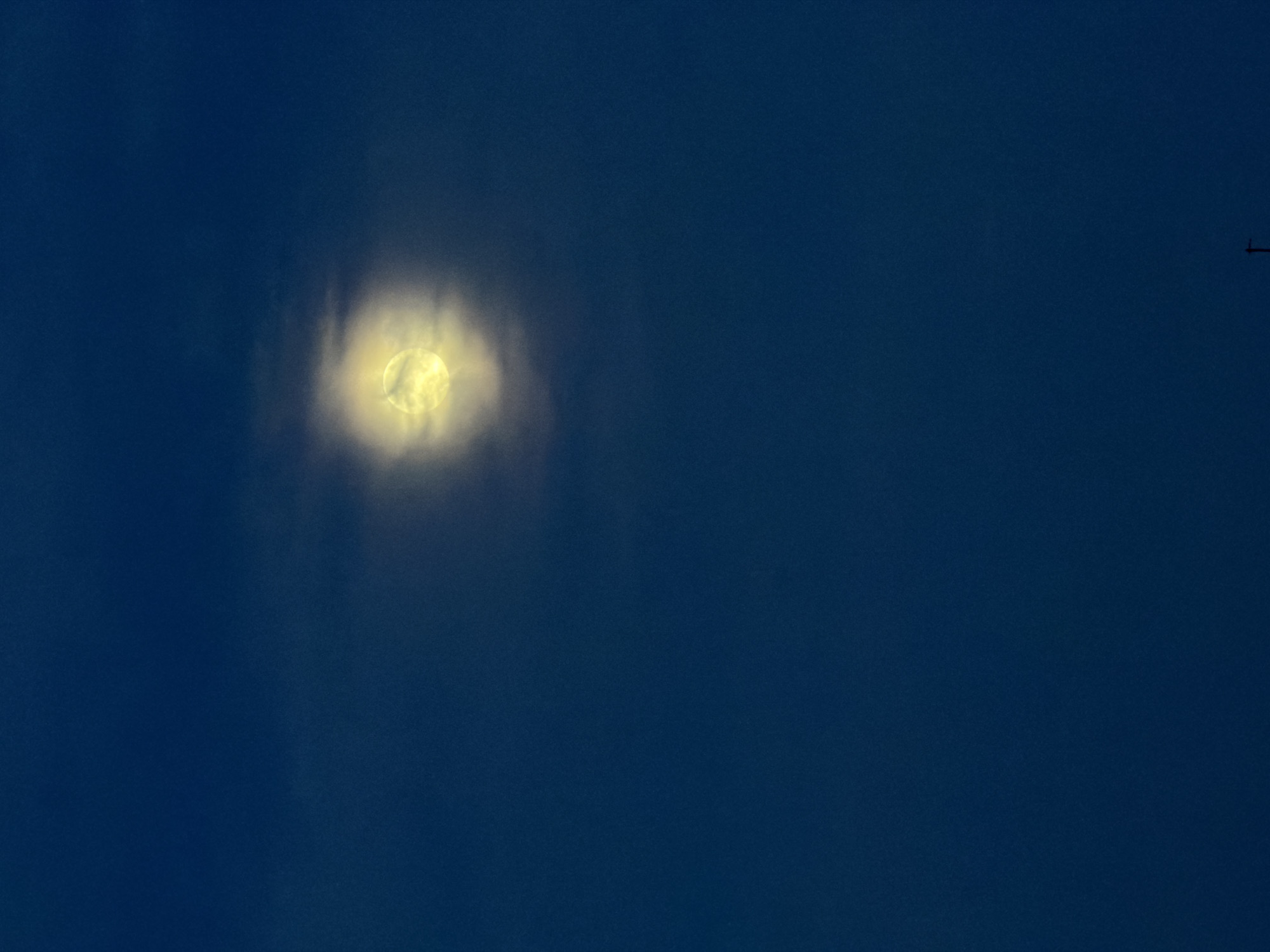 The image displays a night or late twilight sky, featuring a bright moon partially obscured by clouds. The moon, located in the upper left portion of the frame, glows with a strong yellow-white light, its circular form diffused by thin, ethereal clouds passing in front of it, creating a halo effect. The sky is a deep, consistent blue, characteristic of a clear evening or night. In the upper right corner, a small, dark, cross-shaped silhouette is visible against the dark sky, appearing to be a distant aircraft (likely an airplane or helicopter).
The scene suggests a calm night with partial cloud cover. There are no human subjects, specific location cues within the image, or visible text. The overall atmosphere is serene and natural, focusing on the celestial elements. The image is set in Chaikovsky, Russia, according to the provided context.