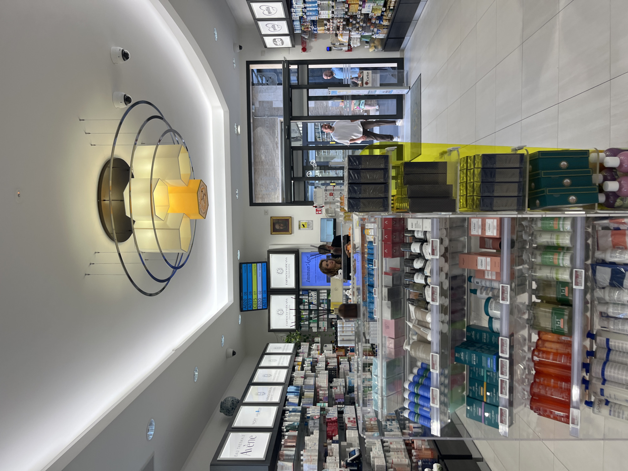 The image depicts the interior of a well-lit pharmacy in Città del Vaticano, Vatican City. The pharmacy features a modern design with white tiled floors and brightly lit white ceilings. Shelving units line the walls, filled with a variety of pharmaceutical and cosmetic products, including bottles, boxes, and tubes.
In the foreground, a display rack showcases numerous beauty and skincare items. These include rows of blue and white bottles, a selection of beige and white creams and lotions, and dark-colored boxes. Further down the rack are green boxed products, and at the bottom, there are rows of purple bottles.
Towards the back of the pharmacy, near the entrance, two men are visible. One man, wearing a white t-shirt and brown pants, stands inside the entrance. Another man, in a blue shirt, is positioned by a counter near the entrance. A female employee, with blonde hair, is also visible behind a counter, interacting with a computer system.
A large digital display board hangs from the ceiling, showing various codes and numbers. A prominent chandelier with multiple hexagonal lamps, some of which emit a yellow light, is suspended from the ceiling. Security cameras are visible mounted on the ceiling in several locations. Posters and framed artwork are displayed on the walls. The entrance to the pharmacy is through a set of glass double doors.