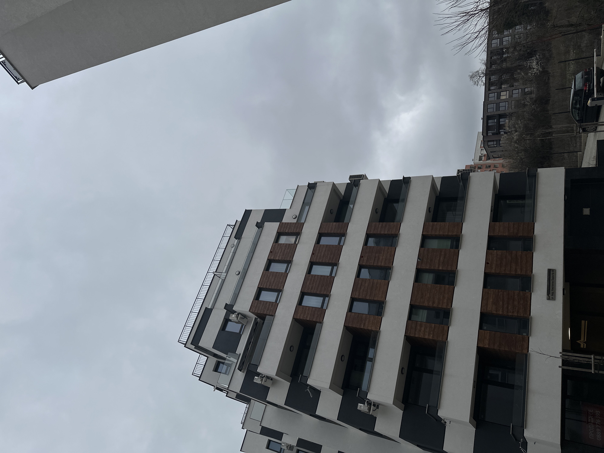 The image presents a low-angle view of a modern, multi-story apartment building in Sofia, Bulgaria, under an overcast sky. The building features a white facade with vertical sections of brown wooden panels and dark, likely black, accents. Windows are rectangular and evenly spaced along the vertical sections. The building's design incorporates staggered, stepped sections, creating protruding balconies. A portion of another building is visible in the upper left corner. Another dark building with multiple windows is visible in the background to the right, with a parked car in front of it. The angle suggests the photo was taken from street level, looking upwards. A sign with Bulgarian writing "Ул. Акад. Капустин Ников No. 44" is visible on the side of the building, and a sign with phone number is visible at the base of the building. Bare tree branches are visible.