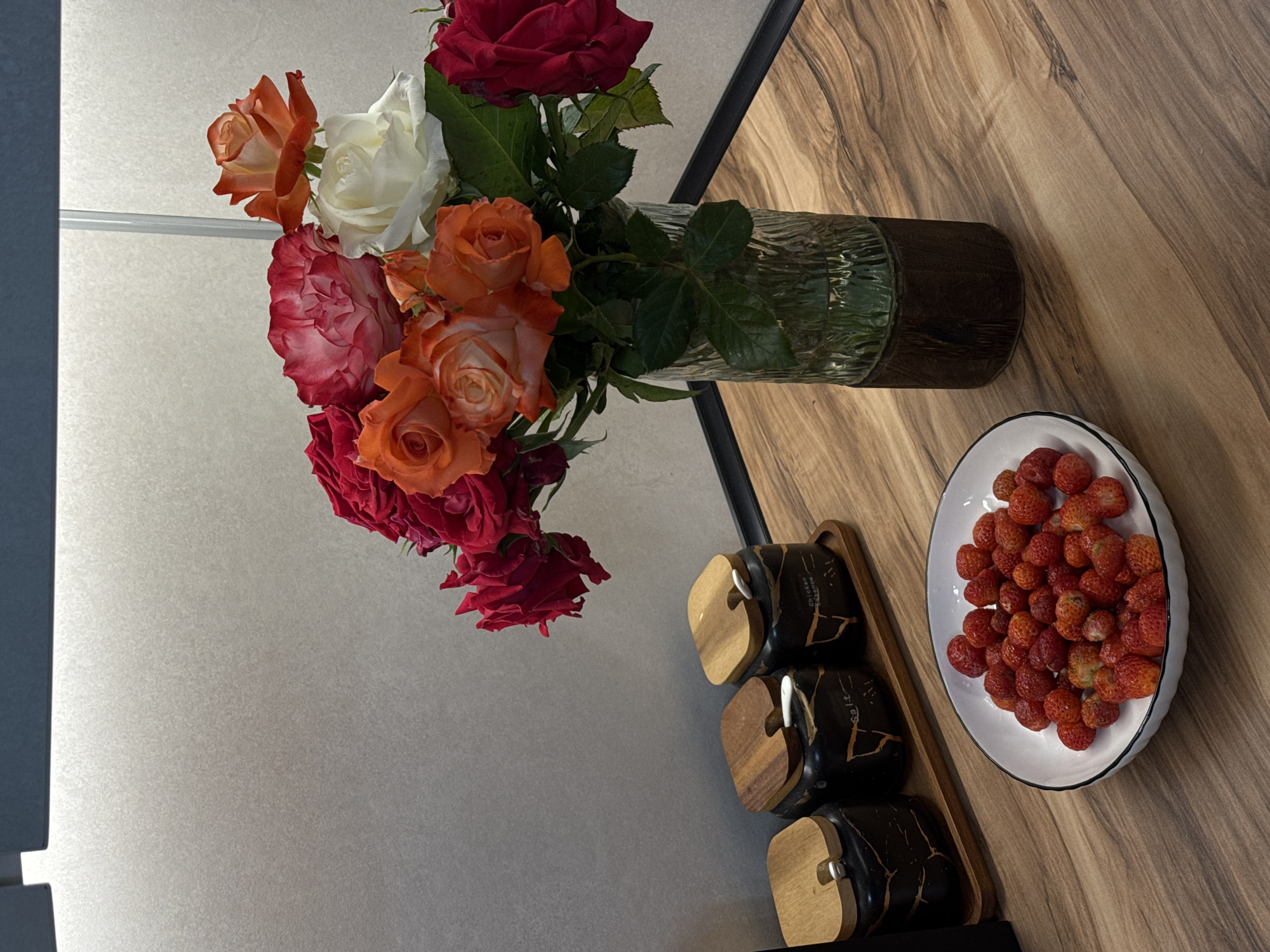 A still life image shows a bouquet of roses in various colors, including red, orange, white, and pink, in a clear glass vase with a dark base. The vase rests on a wooden table. To the left of the vase are three black ceramic jars with wooden lids, arranged on a wooden tray. The jars appear to be spice containers, with one labeled "Salt" and another with "Chicken Essence." In the foreground, a white bowl filled with fresh strawberries is also on the table. The background consists of a textured gray wall. The lighting suggests an indoor setting, possibly a kitchen or dining area. The overall scene is neatly arranged, with a focus on the natural elements of the flowers and fruit. There are no people or animals present in the image. The image does not provide any specific information about the city of Mikhaylovsk, Russia.