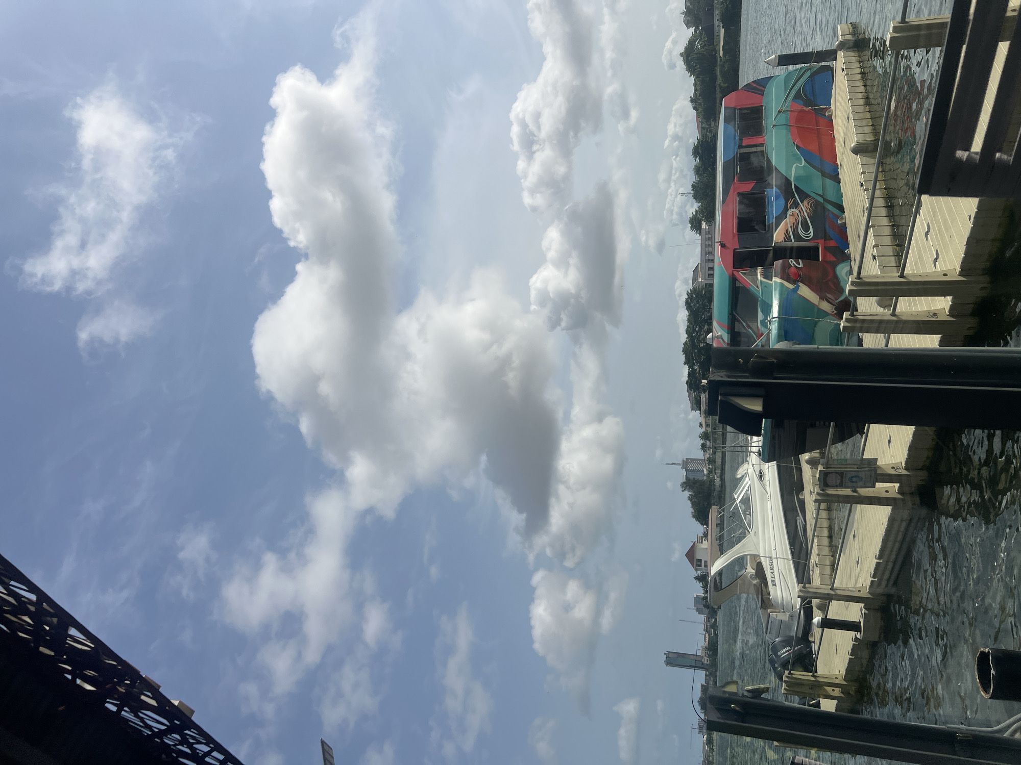 The image depicts a waterfront scene, likely in Ikoyi, Nigeria. The sky is predominantly blue with scattered white clouds. On the right side of the frame, two boats are docked at a wooden pier. One boat is a colorful, multi-decked vessel with artistic designs, while the other is a smaller, white Larson boat with a Yamaha engine. The water is calm, reflecting the sky and surrounding structures. In the lower left corner, a portion of a dark, iron structure, possibly a bridge or building framework, is visible. The scene is captured in daylight.