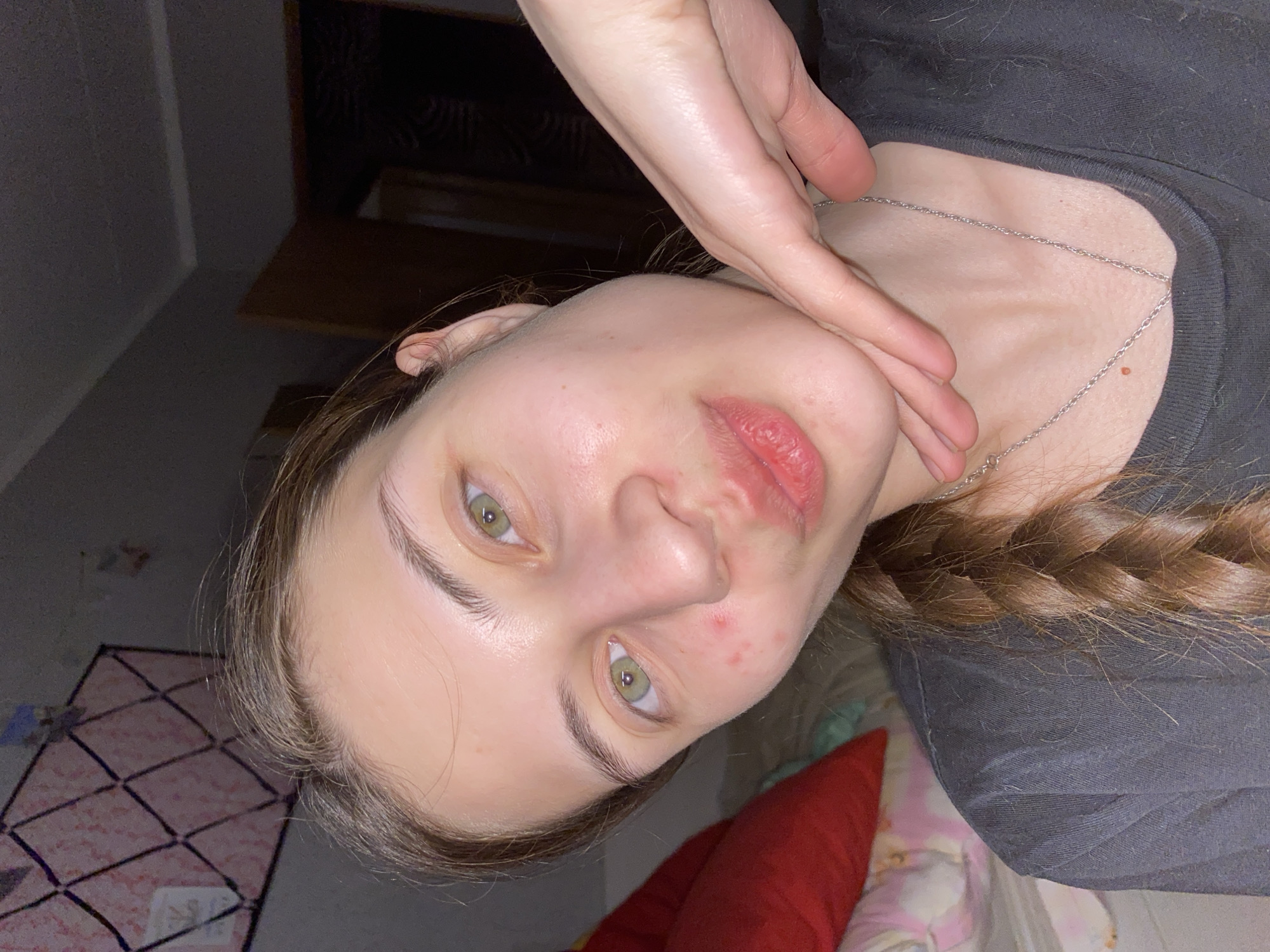 A close-up, tilted portrait features a fair-skinned person with visible skin imperfections. They have green eyes and braided auburn hair. They are wearing a silver necklace and a dark-colored shirt. Their hand is touching their face. The background reveals a bedroom setting with a bed covered in bedding. The location in Votkinsk, Russia is not discernible from the image.