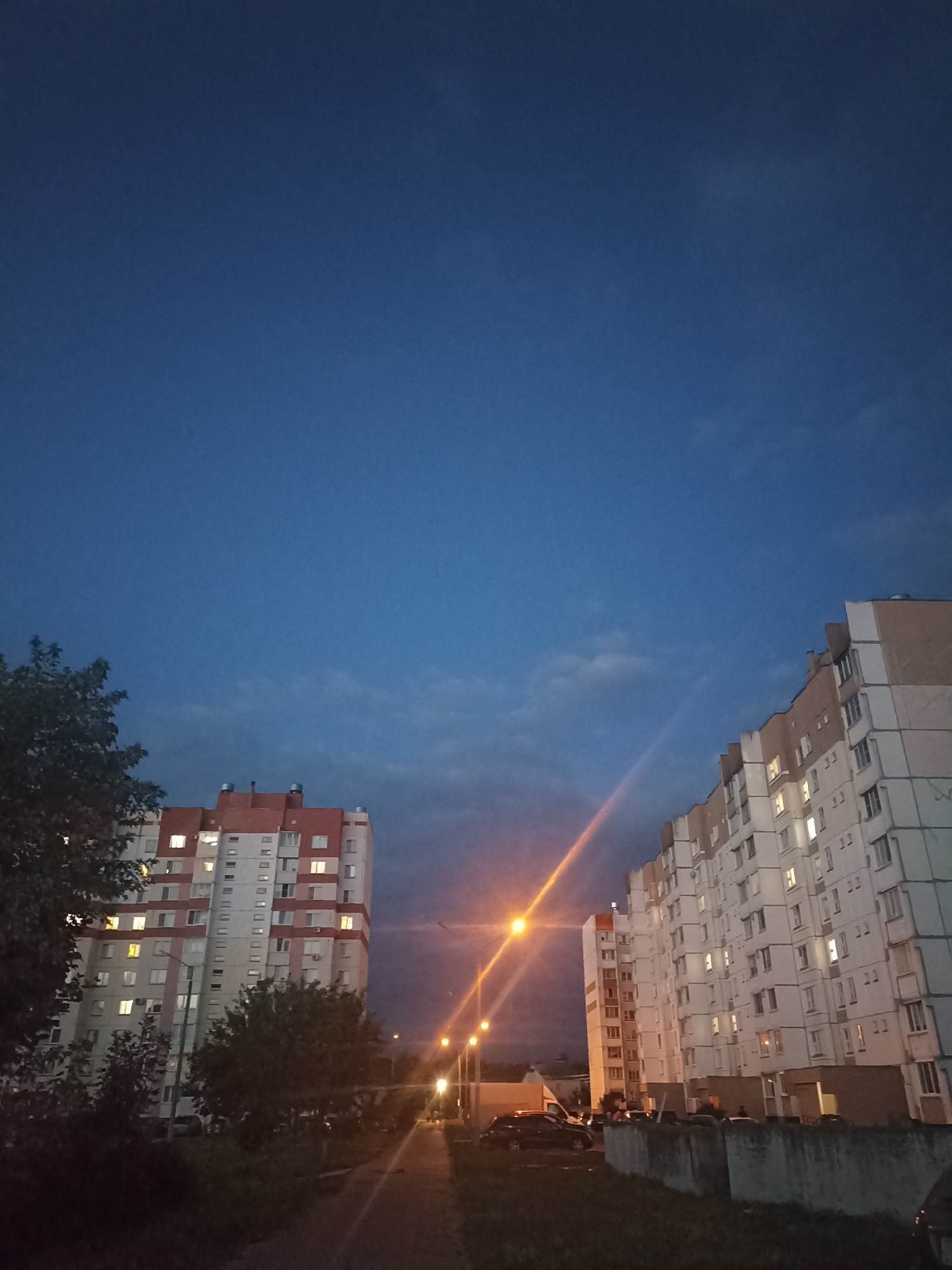 An exterior, eye-level, low-angle, medium shot captures a twilight scene of two apartment buildings in Homyel, Belarus. The sky above is a deep blue, with faint clouds visible. The building on the left is taller and features a red and white facade. The building on the right is wider and has a beige and white exterior. Both buildings have numerous windows, with some illuminated from within, indicating occupancy. A street with streetlights stretches between the buildings, casting an orange glow. Cars are parked along the street and in front of the buildings. Trees are visible in the foreground and between the buildings, some of which are leafy, while others appear bare. The scene is characterized by dim lighting and the transition from day to night.