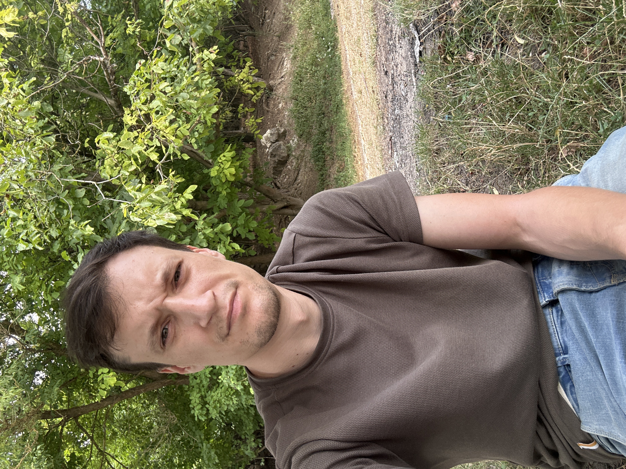 A medium-shot, eye-level, outdoor selfie shows a young adult male sitting in a grassy area with trees in the background. The man, who appears to be in his late twenties, has short brown hair, a light complexion, and is looking directly at the camera with a furrowed brow. He is wearing a brown short-sleeved t-shirt and blue jeans. His right arm is bent and resting on his lap. The background consists of lush green trees and foliage, with a dirt path and dry grass visible in the foreground. The setting is consistent with a park or natural area. The location is identified as the city of Makiv, Ukraine.
