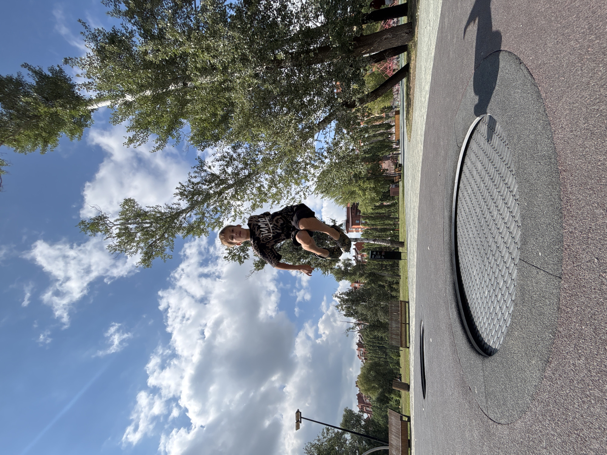 A boy is captured mid-air, mid-jump on an in-ground trampoline at an outdoor recreation area. He is wearing a dark t-shirt and shorts, with camouflage-patterned shoes. The trampoline is circular with a metal mesh surface, embedded within a dark, rubberized ground cover. In the background, several tall, slender trees with green foliage dominate the scene. Beyond the trees, a city skyline is visible, featuring numerous red-tiled roofs and buildings. The sky is bright blue with scattered white clouds. The location is the "Stadion" recreation area in Orenburg, Russia.