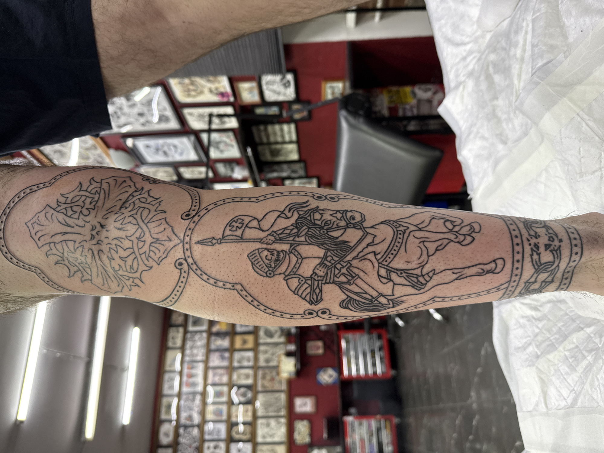 An arm, likely male given the visible hair, is positioned horizontally in the foreground, showcasing a fresh tattoo. The tattoo, rendered in black outline, spans a significant portion of the forearm and features intricate, medieval-inspired designs. The central and most prominent part of the tattoo depicts a dynamic jousting scene with two armored figures on horseback, one seemingly striking the other with a spear. This scene is enclosed within an ornate, scalloped border. To the left of the jousting scene, another section of the tattoo displays a complex, stylized cross or floral pattern, also framed by the decorative border. To the right, a smaller band features a geometric or knotwork design.
The setting appears to be a tattoo studio. In the background, out of focus, are red walls adorned with numerous framed pictures or tattoo flash designs, suggesting various potential tattoo patterns. Bright, vertical strip lights illuminate the room. On the right side of the image, a crinkled white sheet or paper material, possibly a protective cover or medical wipe, is visible. Another piece of white paper with tape is also discernible. No discernible text is visible on the tattoo or in the background. The image captures the fresh application of the tattoo, likely taken shortly after completion in the studio.