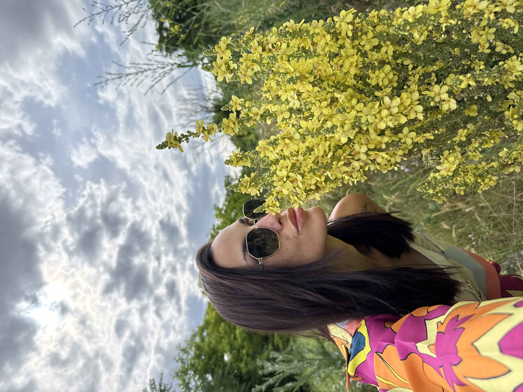 In a field with tall green grass and trees in the background, a woman with long dark hair and sunglasses leans towards a large cluster of bright yellow flowers. She is wearing a white top and a colorful shirt with orange, pink, and yellow floral patterns. The sky above is cloudy with patches of sunlight breaking through. The scene is outdoors, with vegetation indicating a natural environment. The image composition focuses on the woman and the flowers, suggesting an interaction with nature. The presence of clouds and sunlight indicates it is daytime. The overall mood is relaxed and scenic.