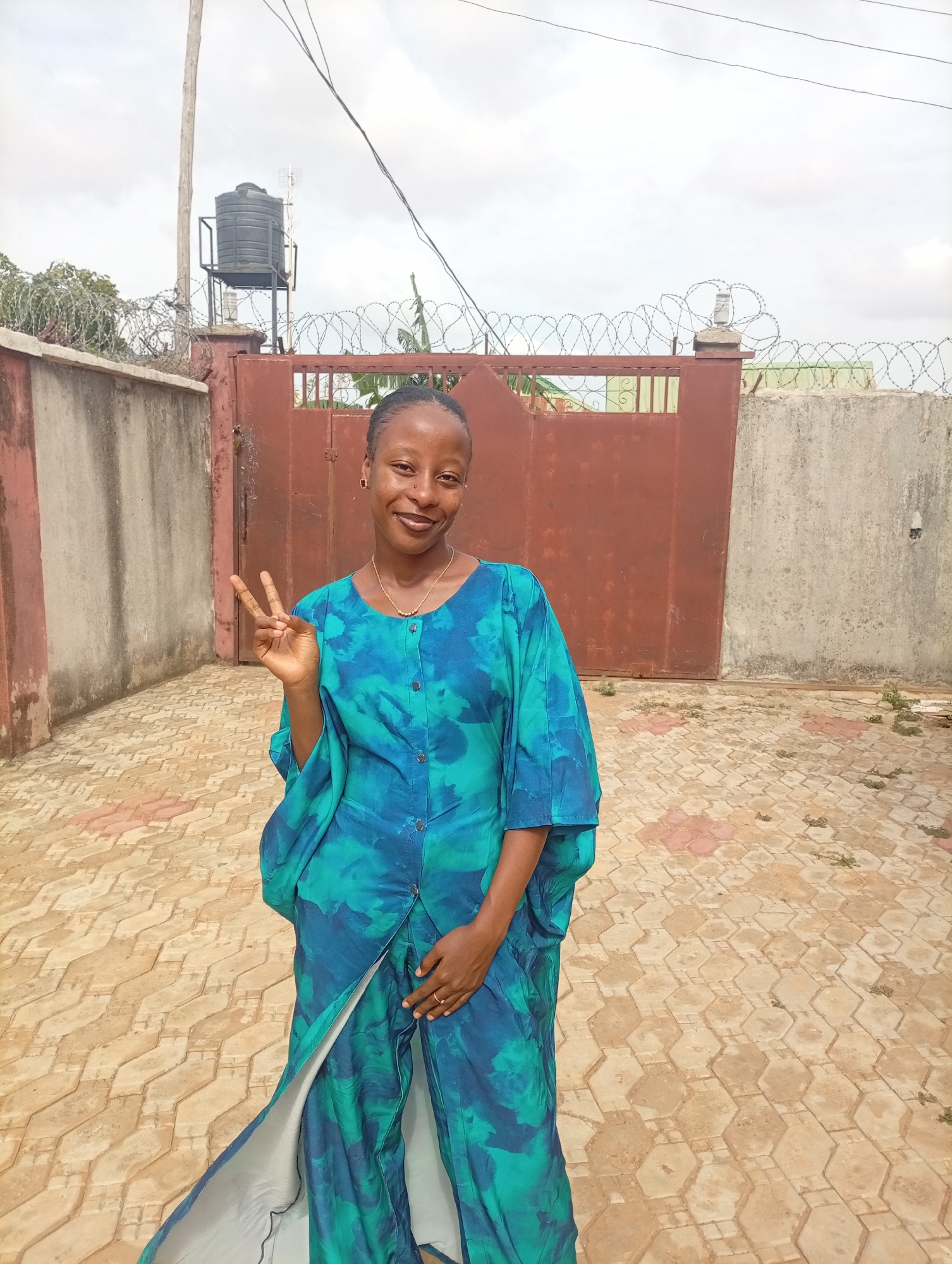 A young, brown-skinned woman is standing outside, posing for a photo with a peace sign. She has her hair pulled back, is wearing a blue and green tie-dye style outfit and gold jewelry. She is standing on a tan tiled area. Behind her is a tall metal gate, concrete walls topped with barbed wire, and a black water tank on a platform. The image was taken in Rido, Nigeria.