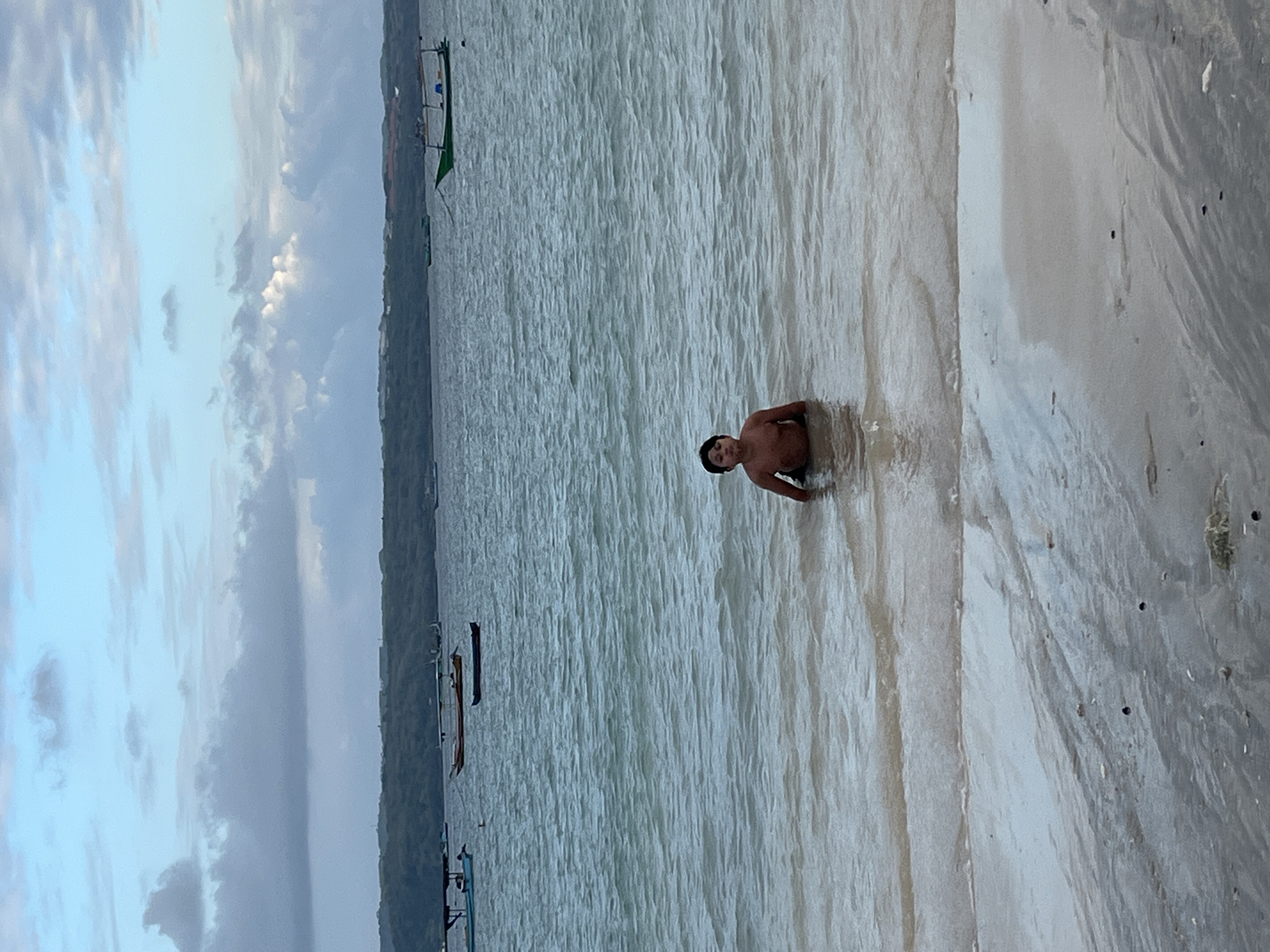 A young boy is standing in the shallow water near a beach. He is partially submerged, with the water coming up to his chest. The boy has dark hair and is facing the camera. The background shows a body of water with several boats anchored. In the distance, there is a lush green coastline with a few buildings visible. The sky above is a mix of blue and gray clouds, suggesting it might be early morning or late afternoon. The sand on the beach in the foreground is wet and has a light brown hue with some darker specks. The overall scene depicts a serene coastal environment in Kuta, Indonesia.