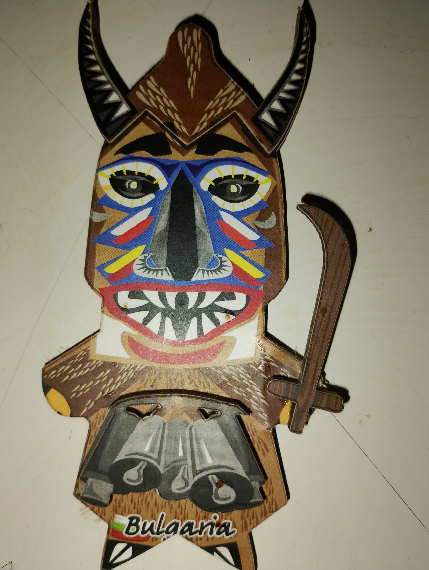 A close-up, overhead shot captures a colorful, cardboard cutout of a traditional Bulgarian Kukeri mask. The mask depicts a stylized, monstrous face with prominent black horns adorned with a zigzag pattern in black, white, and grey. Large, expressive eyes are outlined in blue and yellow, with red and yellow accents around them. The nose is black, and a wide mouth reveals sharp, white teeth against a red background. The mask has a brown, textured pattern resembling fur on its cheeks and lower chin. Attached to its right side is a wooden cutout of a sword. Below the mask, a skirt-like element features three grey bells. The bottom of the cutout displays the word "Bulgaria" in white lettering, with a small Bulgarian flag to its left. The cutout is placed on a light-colored, flat surface, possibly a table, with faint lines suggesting creases or seams in the surface. The lighting creates subtle shadows around the cutout. The background is a plain, light-colored surface.