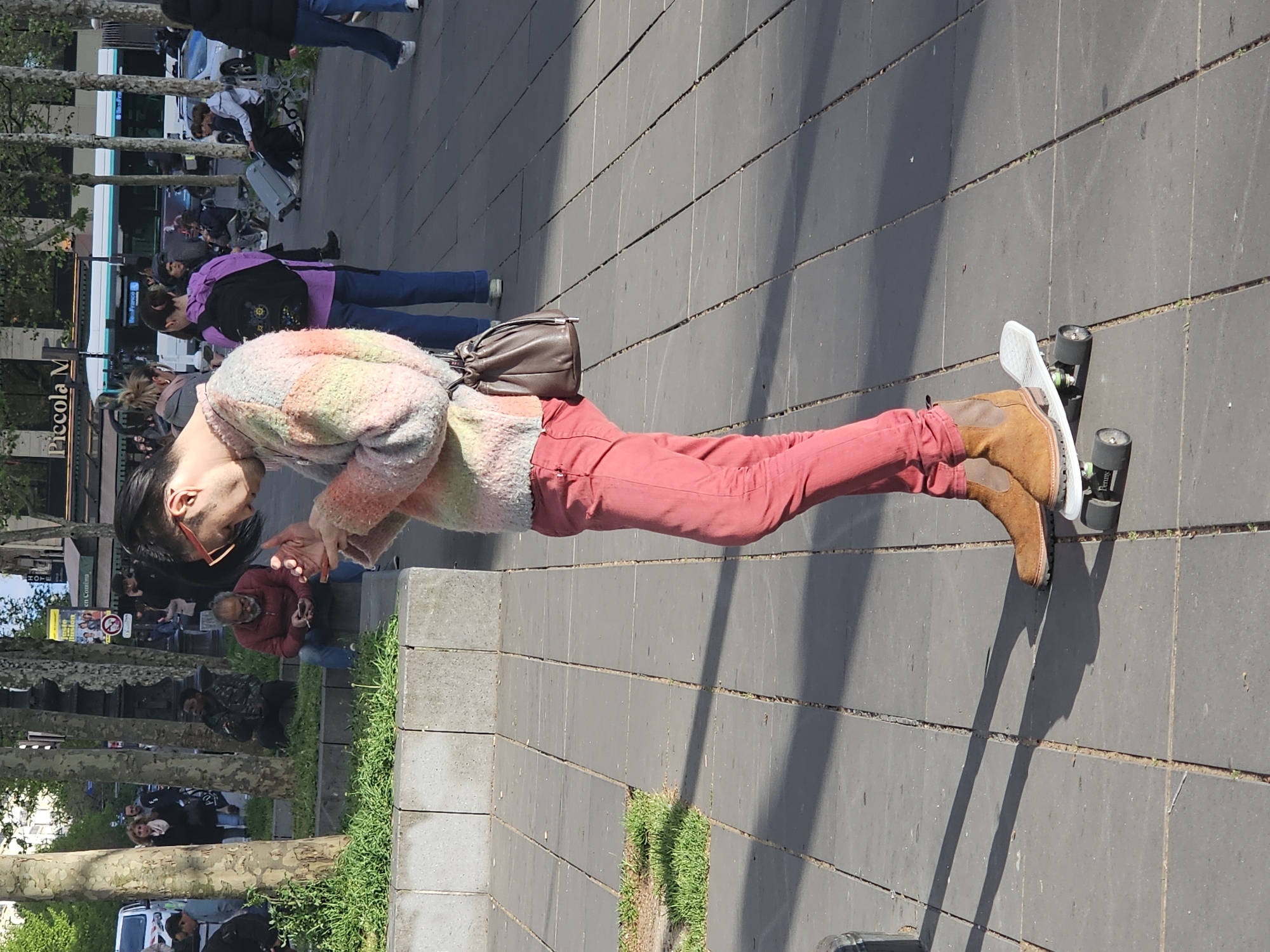 A medium shot shows a person riding a skateboard on a sidewalk on a sunny day in Paris, France. The person is wearing red pants, brown boots, and a multi-colored sweater. They have short dark hair and are wearing red glasses. They also have a brown purse slung over their shoulder. The skateboard is white with black wheels. The sidewalk is made of gray rectangular pavers. Behind the person are trees and other people walking along the sidewalk. There is a bus in the background.