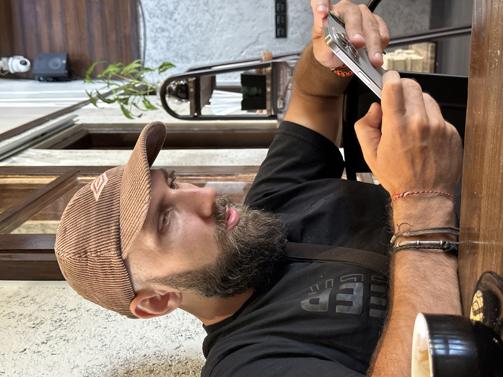 A medium shot captures a man wearing a brown corduroy baseball cap and a black t-shirt, seated at a wooden table in what appears to be a cafe. He has a full beard and is intently looking at his smartphone, which he holds in his right hand. His left hand rests on the table near a black coffee cup. The background shows a textured wall, wooden elements, and a shelf with plants and other items. The overall lighting suggests an indoor environment, likely during the day. The man has several bracelets on his wrists, including a red and white string bracelet.
