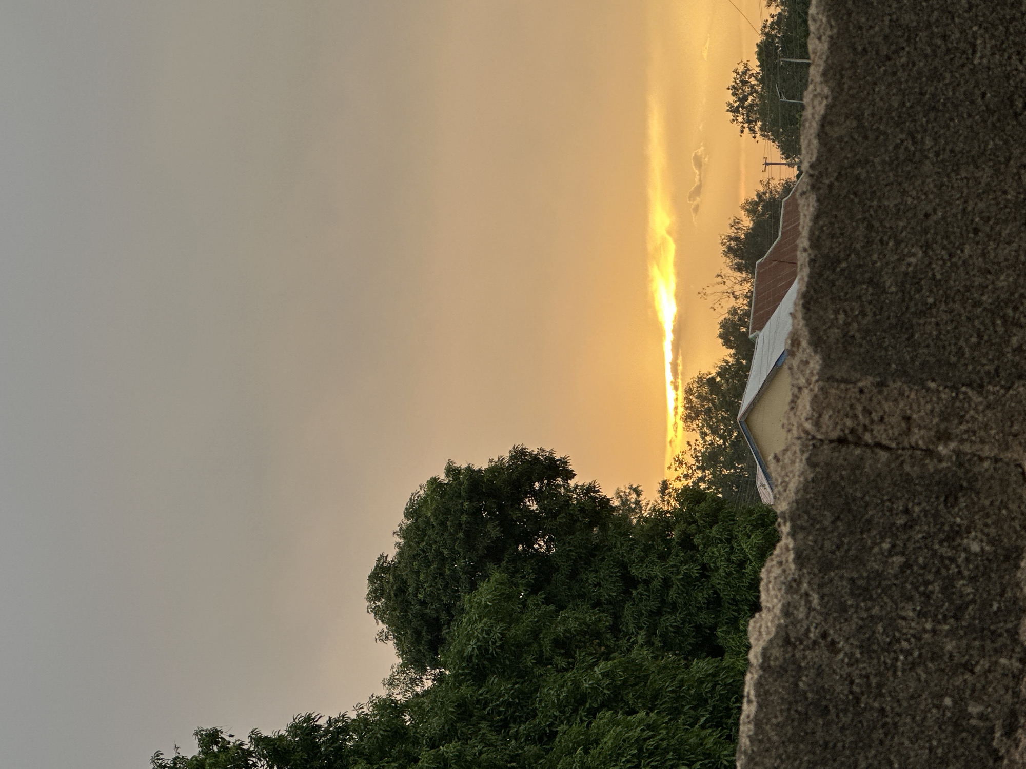 The image captures a sunset scene in Bukuru, Nigeria. A large, green tree dominates the lower portion of the frame, contrasted against a greyish sky transitioning to a bright, yellow-orange sunset directly overhead. A streak of bright light is visible through a gap in the clouds. In the upper right corner, a portion of a building roof and trees are visible. The right side of the image is occupied by a rough, grey concrete structure, likely a wall or part of a building, with a prominent vertical crack. No people are visible.