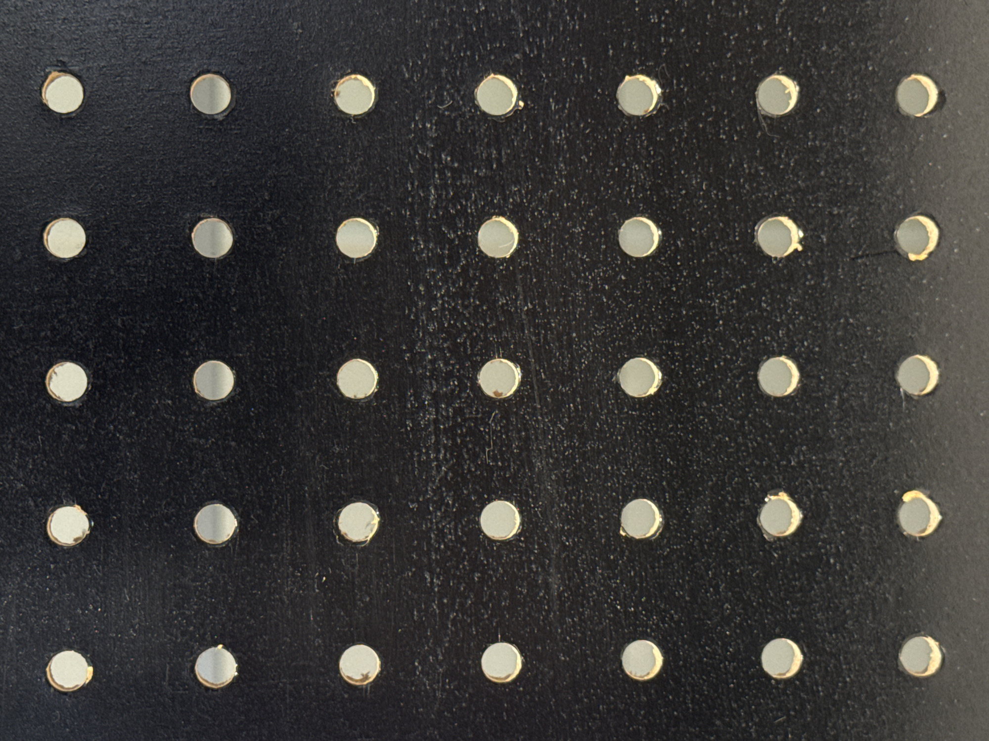 The image is a close-up, top-down view of a black surface with a grid of evenly spaced circular perforations. The perforations appear to be holes through the material, revealing a lighter color, possibly metal, on the underside. The surface itself has a slightly textured, matte finish, with some minor imperfections and dust particles visible. The lighting suggests a diffuse light source, casting soft shadows within the perforations, highlighting their depth. There are no people, animals, or any other discernible objects or actions in the image. The scene is abstract and lacks any specific context related to a location in Moscow, Russia, or any identifiable city or environment.