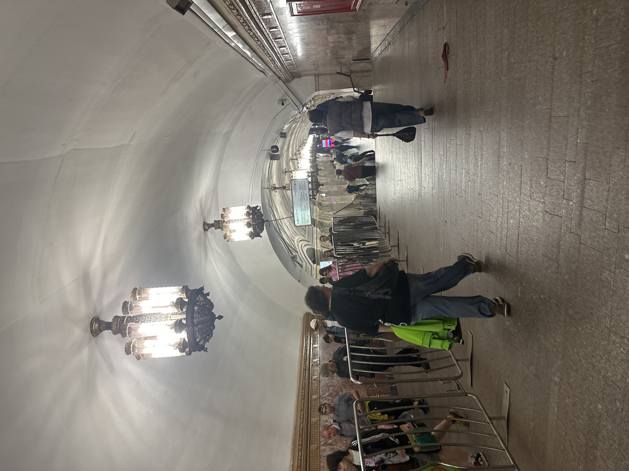 The image depicts a wide, ornate subway station with a high, arched ceiling and elaborate chandeliers. The station is illuminated by the warm glow of these chandeliers, casting intricate shadows on the white walls. The floor is paved with large, light-colored tiles. Several people are visible, most of whom appear to be waiting or walking along the platform. Some are standing behind metal railings, suggesting a queuing area.
One individual in the foreground, facing away from the camera, is wearing blue pants and a black top, carrying a backpack. Another person further down the platform, also facing away, is wearing a grey vest over a lighter shirt and dark pants, holding what appears to be a large, dark object, possibly a paddle or a musical instrument. In the background, more figures are scattered along the platform, some in groups and others alone.
The overall atmosphere seems calm, with no indication of urgency or distress. The lighting and architectural style suggest an indoor, possibly underground, public transport hub. There are no obvious indications of the time of day or weather conditions, as the scene is entirely internal. Some signs are visible in the background, but the text is not legible. The scene appears to be a typical day in a busy subway station.