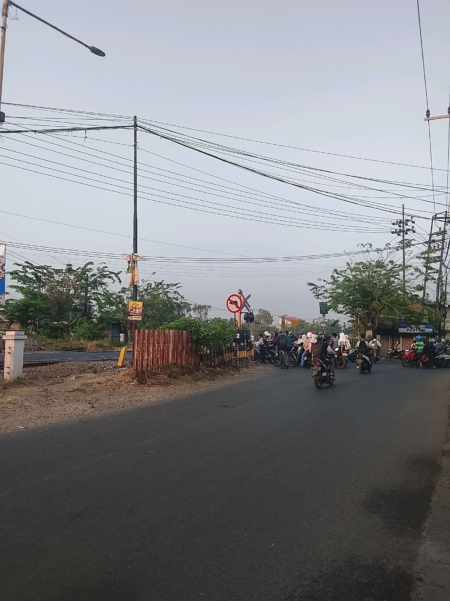 The image depicts a street scene in Bebekan, Indonesia, likely at a railway crossing. A moderate amount of traffic, primarily motorcycles, is stopped at the crossing, suggesting the presence of an approaching train or a closed barrier. The sky is overcast, and the lighting suggests it is either early morning or late afternoon.
Key elements in the scene include:
* **Roadway:** A paved road occupies the foreground and extends into the distance. It appears to be a two-lane road.
* **Traffic:** Numerous motorcycles are parked on the road, with riders waiting. Some are single-rider, while others have passengers. A few motorcycles are in motion.
* **Railway Crossing:** A railway track runs horizontally across the image, intersecting the road. A railway crossing sign, depicting a red circle with a diagonal line and an arrow indicating a turn, is visible on the left side. There is also a yellow marker with the number "2" on it, likely indicating the distance to the crossing or a track number.
* **Vegetation:** Trees and bushes are present on both sides of the road and beyond the railway tracks, creating a somewhat rural or suburban atmosphere.
* **Infrastructure:** Utility poles with numerous overhead wires are prominent in the scene, indicating the presence of electricity and telecommunication lines. A street lamp is also visible.
* **Structures:** A small building with the word "PARFUM" visible on its facade is located on the right side of the road in the background. A rusted metal fence or barrier is present near the railway tracks on the left.
The overall impression is one of a common occurrence at a busy intersection in a moderately populated area, where traffic flow is temporarily interrupted by a railway passage. The scene is composed from a low-angle perspective, likely from a vehicle or standing at street level.