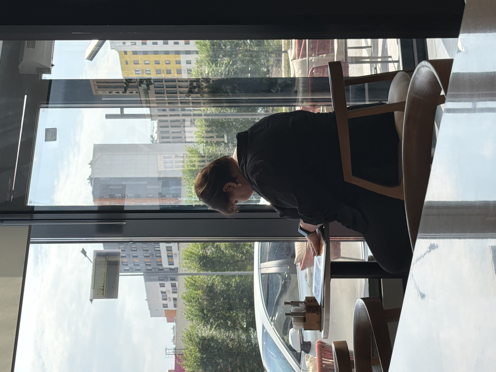 A woman is seated at a table in a cafe in Astana, Kazakhstan, facing a window. She is wearing a black top and appears to be reading a document or menu. A white car is parked outside the cafe, visible through the window. The cafe interior includes wooden chairs and tables, with a reflection of the cafe and the outside environment visible on a glossy tabletop in the foreground. The background shows modern buildings and trees, characteristic of an urban setting.