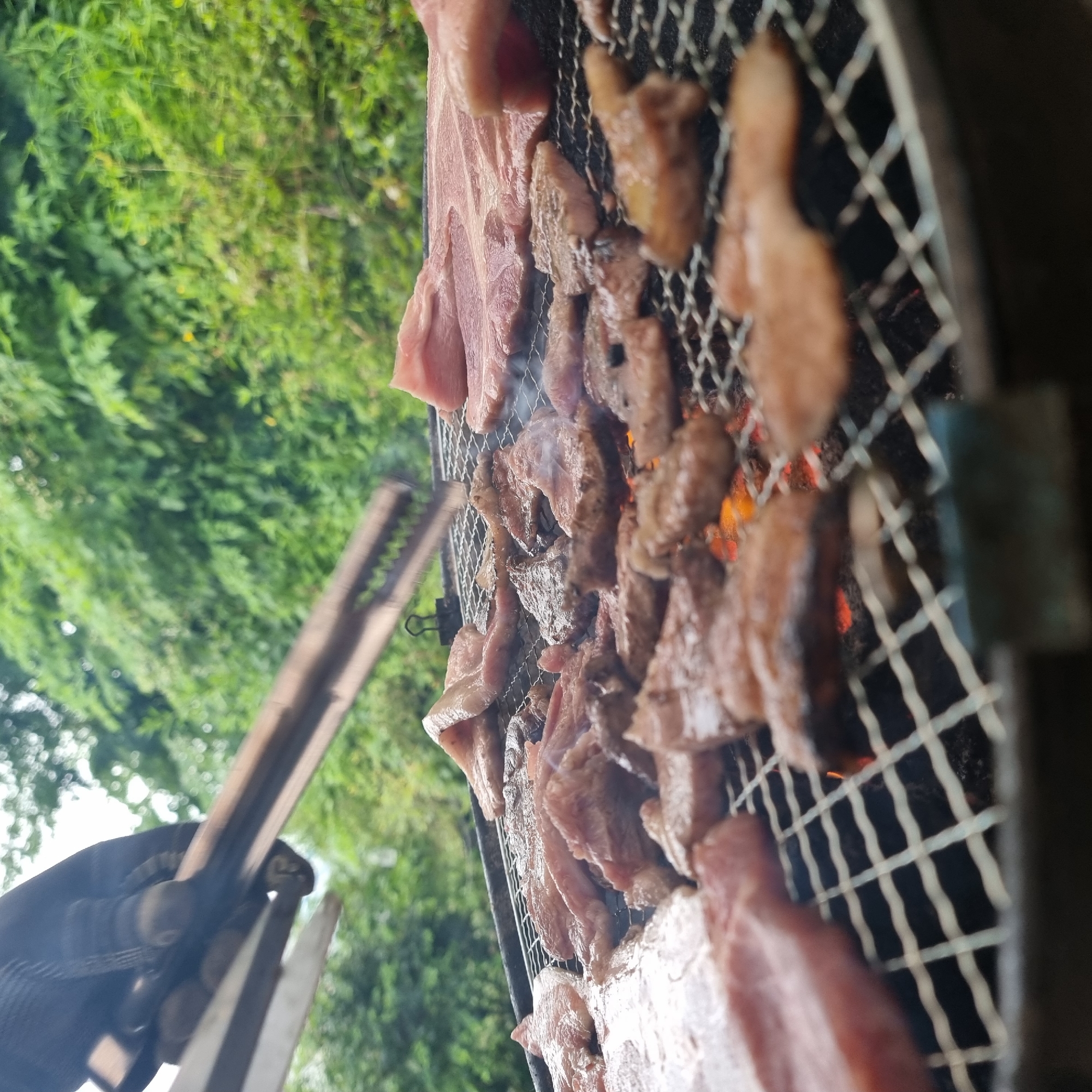 In Dunnae-myeon, South Korea, a close-up, eye-level shot captures an outdoor barbecue scene. A gloved hand grips a pair of metal tongs, positioned above a wire grill laden with numerous slices of raw meat. The meat, predominantly pinkish-red with some marbling, is arranged in layers across the grill. Visible beneath the meat, glowing red embers suggest the grill is actively heated. The background is a blur of lush green foliage, indicating a natural, possibly forested, environment. The lighting appears natural, consistent with daylight.
