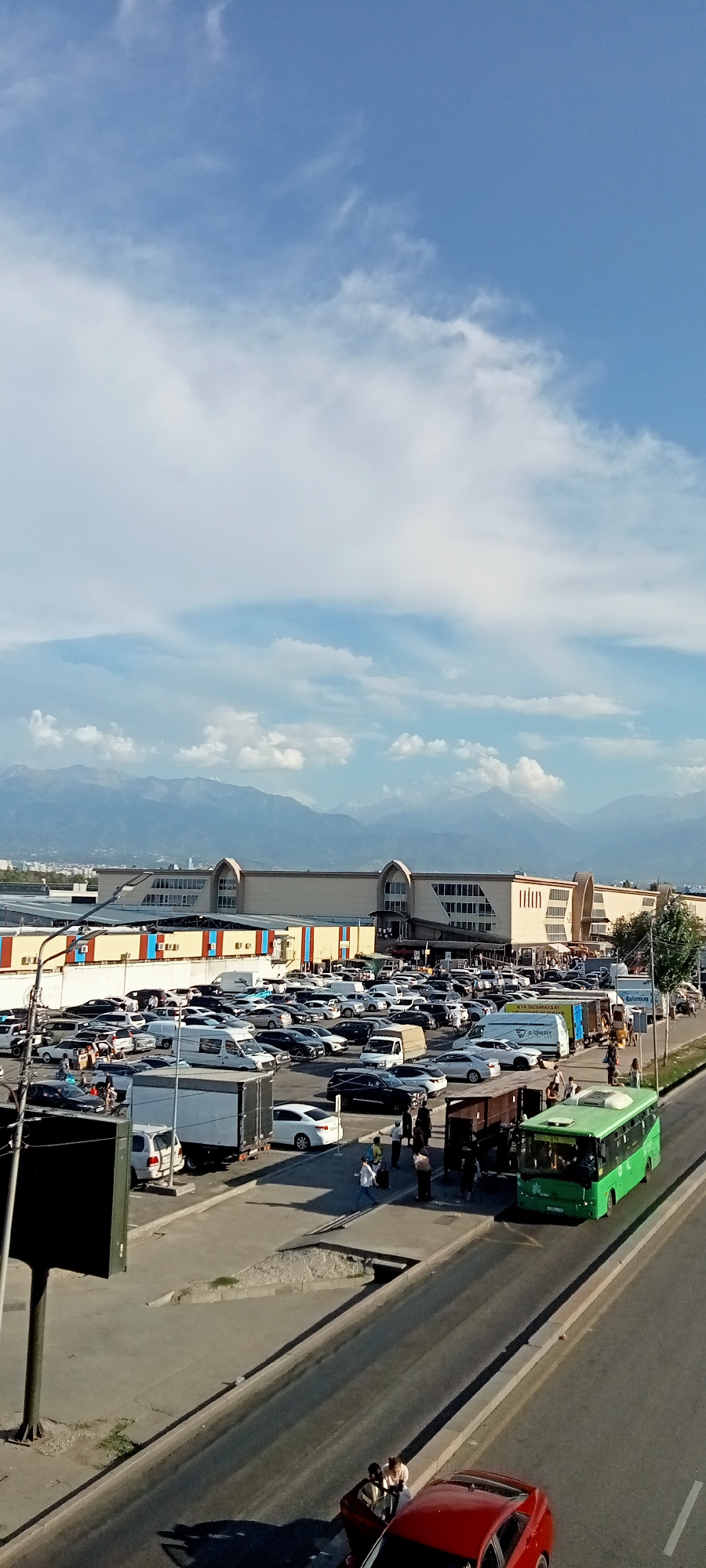 The image captures a wide-angle view of a busy outdoor scene in Almaty, Kazakhstan. In the foreground, a paved road slopes diagonally from the bottom right, with a red car with its door open and two occupants visible. Further up the road, a green bus, identified by signage as "210", is traveling to the right. To the left of the road, a large, paved area is filled with numerous parked cars of various makes and models, suggesting a market or commercial area. Several people are seen walking around the parked vehicles, some with luggage, indicating a potentially transient population or shoppers.
Dominating the mid-ground is a long, multi-story building with a beige facade and dark-tinted windows. The building appears to be a commercial establishment, possibly a shopping center or marketplace, with arched architectural elements visible. Smaller structures, some appearing to be stalls or service areas, are situated in front of the main building and between the parking lot and the road.
In the background, a range of majestic mountains, possibly the Tian Shan range, forms a dramatic backdrop against a bright blue sky dotted with scattered white clouds. The overall impression is one of a bustling urban environment set against a stunning natural landscape. The lighting suggests it is daytime, likely late afternoon given the long shadows cast by the objects.