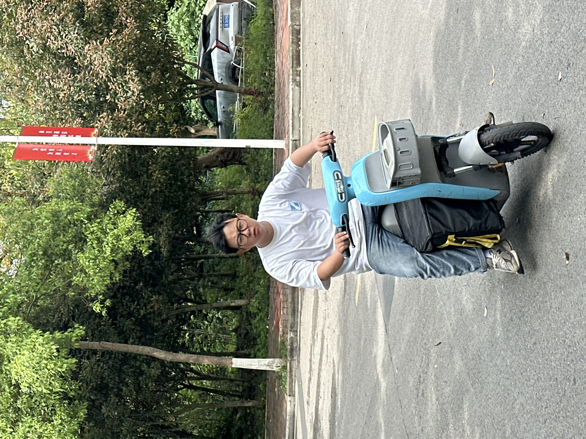 A person wearing glasses, a white shirt, and blue jeans is standing beside a blue electric scooter on a paved surface. The person is facing the camera and has their hand on the scooter's handlebar. Trees and a road sign are visible in the background, and a car is parked further back. The location is the city of Jiangjin, China.