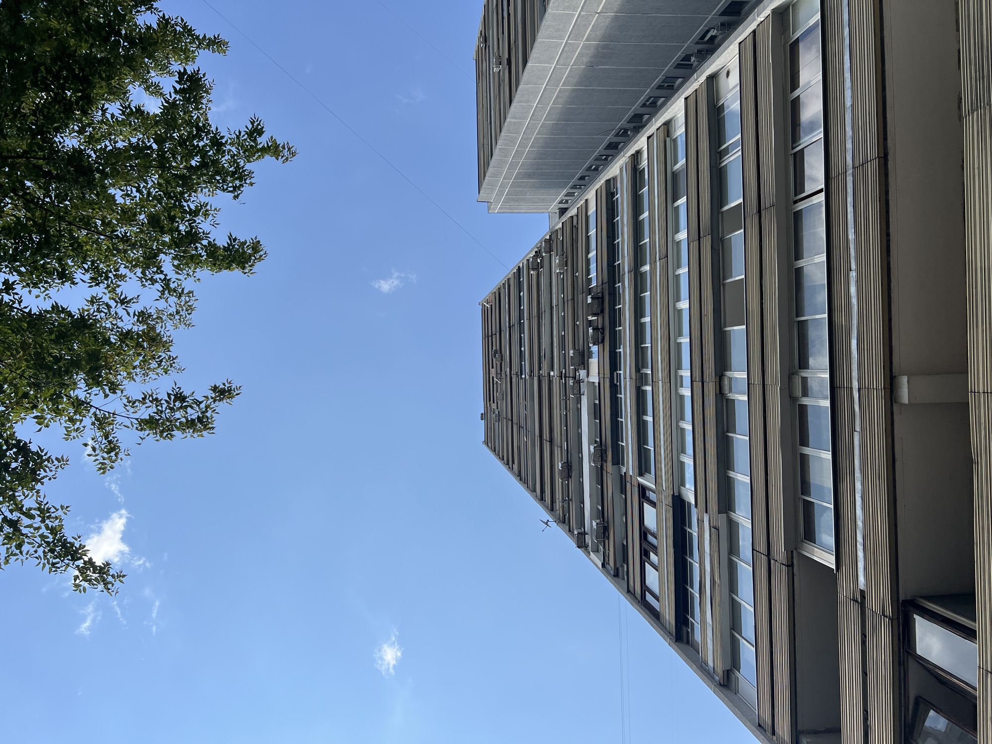 A low-angle, worm's-eye view of a brutalist apartment building in Moscow, Russia, is captured under a clear blue sky dotted with a few wispy clouds. The facade of the building is a grid of windows and horizontal balconies, with many of the balconies featuring external air conditioning units. To the right of the primary building, another similar structure is partially visible, its grey concrete facade also marked by a grid of windows. The branches of a leafy green tree overhang the top of the frame, providing a natural contrast to the rigid lines of the architecture. Electrical wires stretch across the sky, adding to the urban atmosphere. The image is devoid of any human presence or activity.