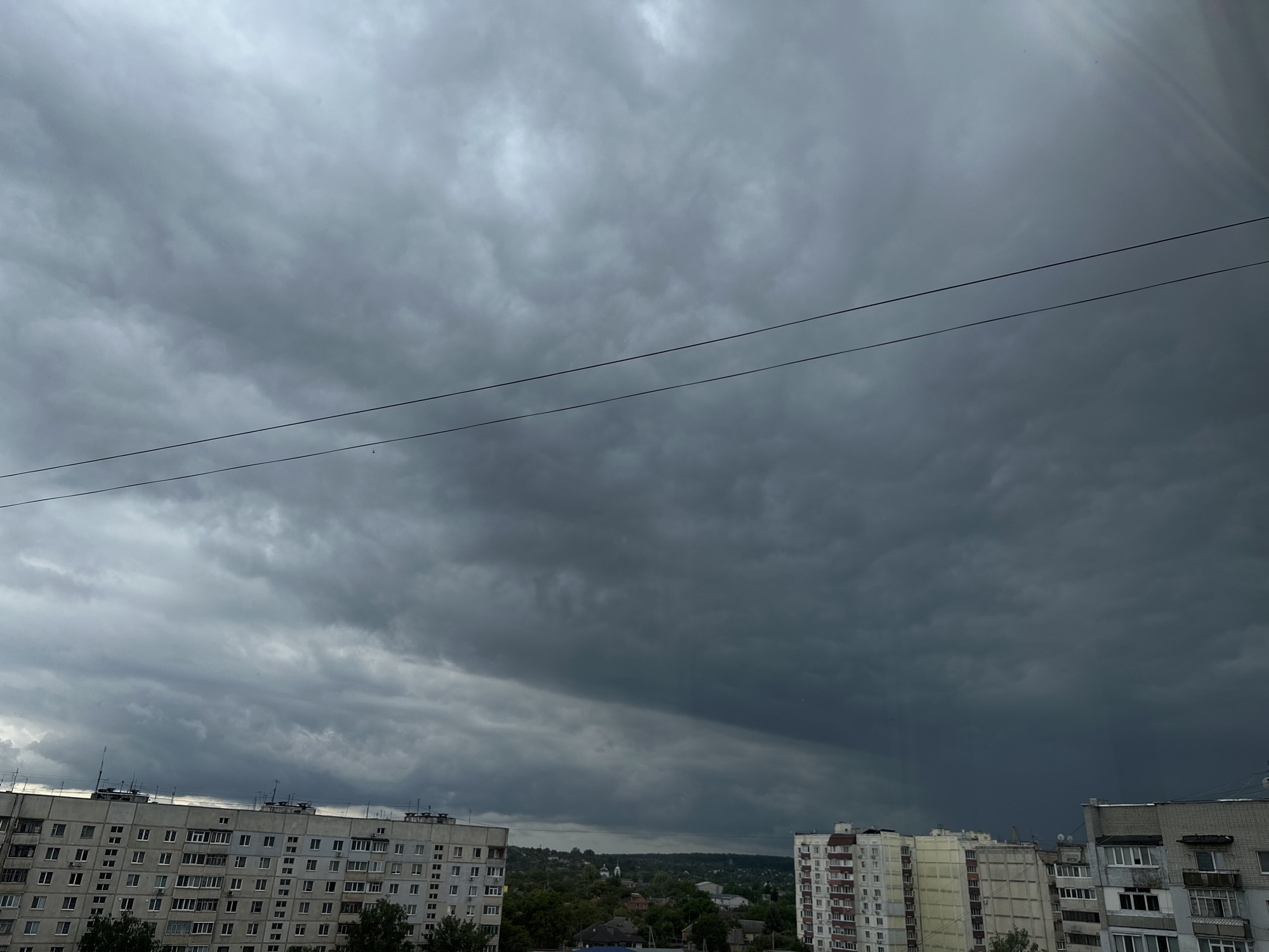 The image captures a view of the sky over a city with several multi-story buildings. The sky is heavily clouded with dark, ominous-looking clouds, suggesting the possibility of rain or a storm. Two dark wires are visible stretched across the upper portion of the frame. In the foreground, the tops of several apartment buildings are visible. The buildings are characterized by their rectangular shapes, numerous windows, and balconies. Beyond the buildings, a landscape with trees and some smaller structures can be seen, with a faint hint of a building with a dome in the distance. The overall impression is one of a city preparing for or experiencing inclement weather.