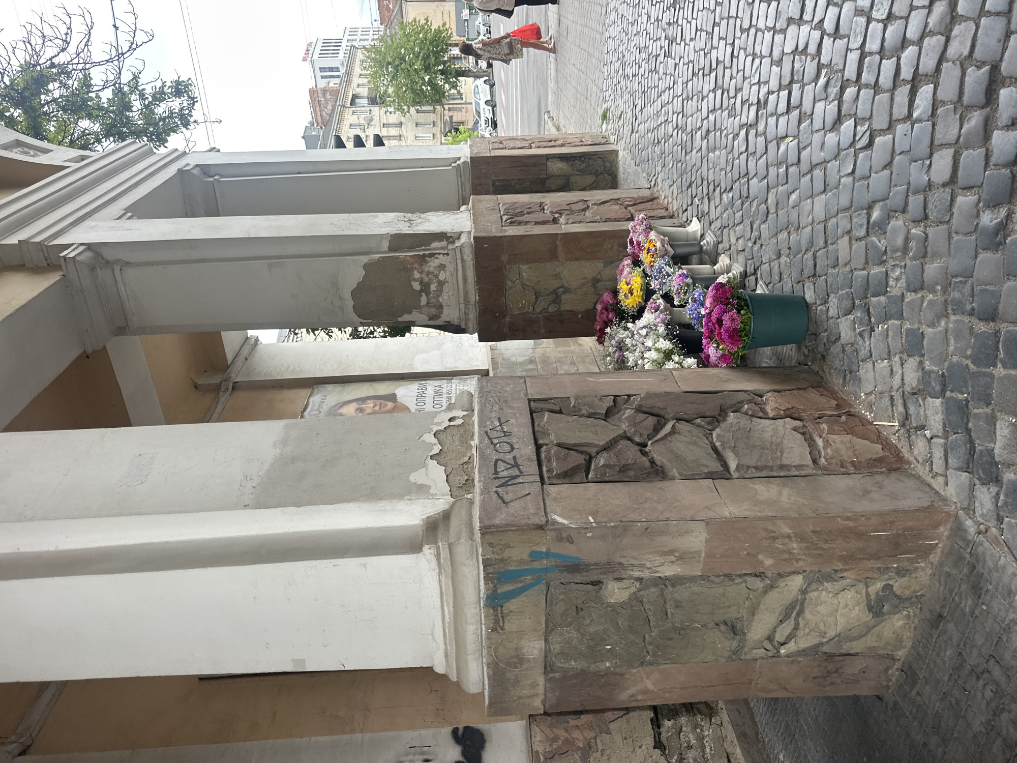 The image shows a street scene in Ternopil, Ukraine, near the Ternopil Regional State Administration building. In the foreground, a collection of flower buckets is arranged on a cobblestone sidewalk. The flowers are a vibrant mix of colors, including pink, yellow, purple, and white. Behind the flowers, a stone-clad column of a building is visible, with graffiti on its surface. Further in the background, several people are walking on the sidewalk, and cars are visible on the street. The architecture suggests a mix of older and more modern buildings. The sky is overcast, indicating a cloudy day.