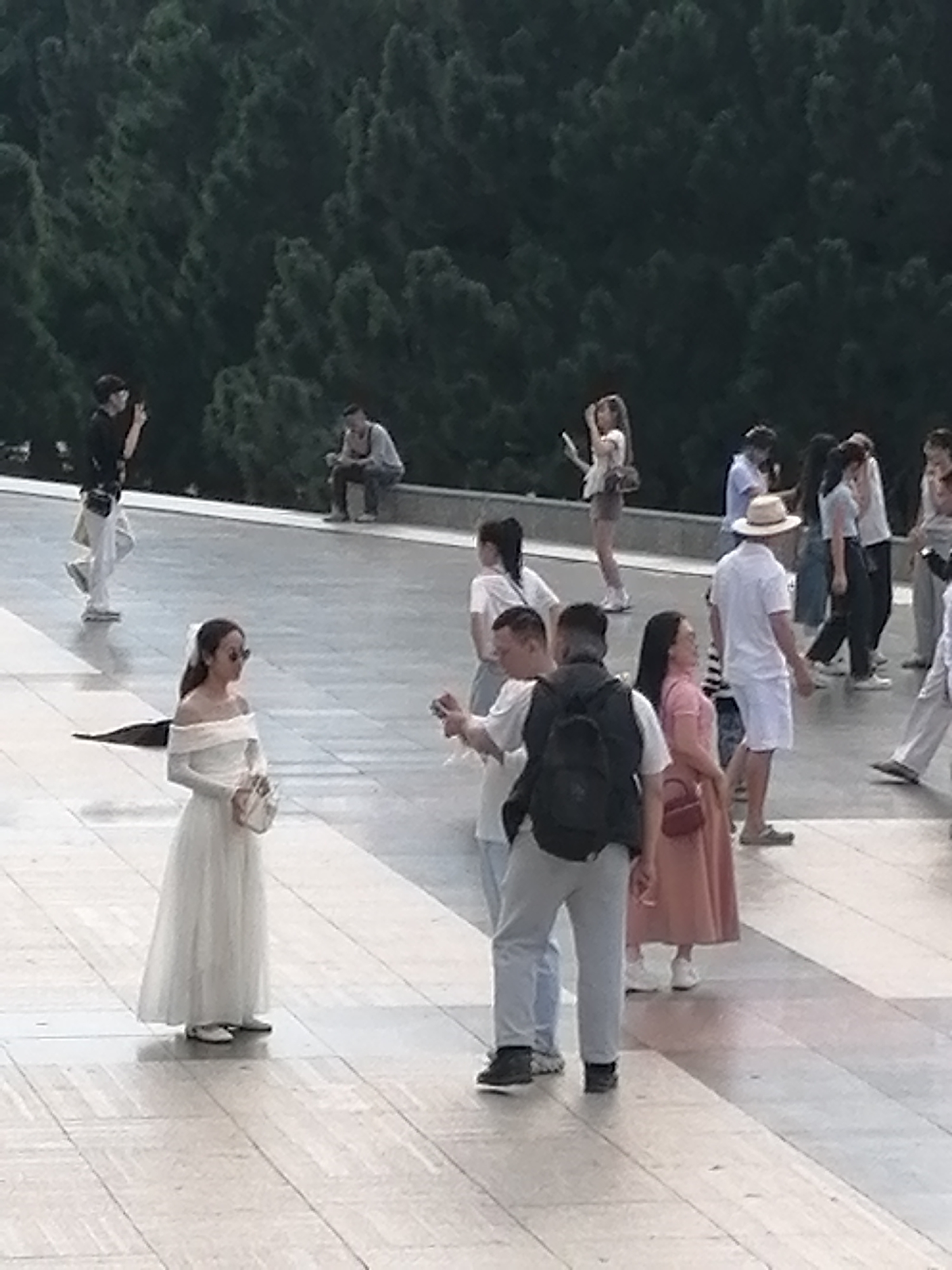 The image depicts a group of people in what appears to be a public outdoor space with a treed background. In the foreground, a woman in a white, off-the-shoulder dress and sunglasses stands facing left, holding a light-colored purse. To her right, a man wearing a dark backpack and gray pants faces away from the camera. To the left of the woman in white, another woman in a dark top and light pants walks away from the camera, holding a phone. In the middle ground, several other individuals are visible, including a man in a white shirt and hat, and others in casual attire. In the background, a man is seated on a ledge. The ground is paved with large, light-colored tiles. The presence of a similar tiled pathway and the lush greenery suggest this could be a location like the Dinh 3 (Bao Dai Palace) in Da Lat, Vietnam, which features such architecture and landscaping.