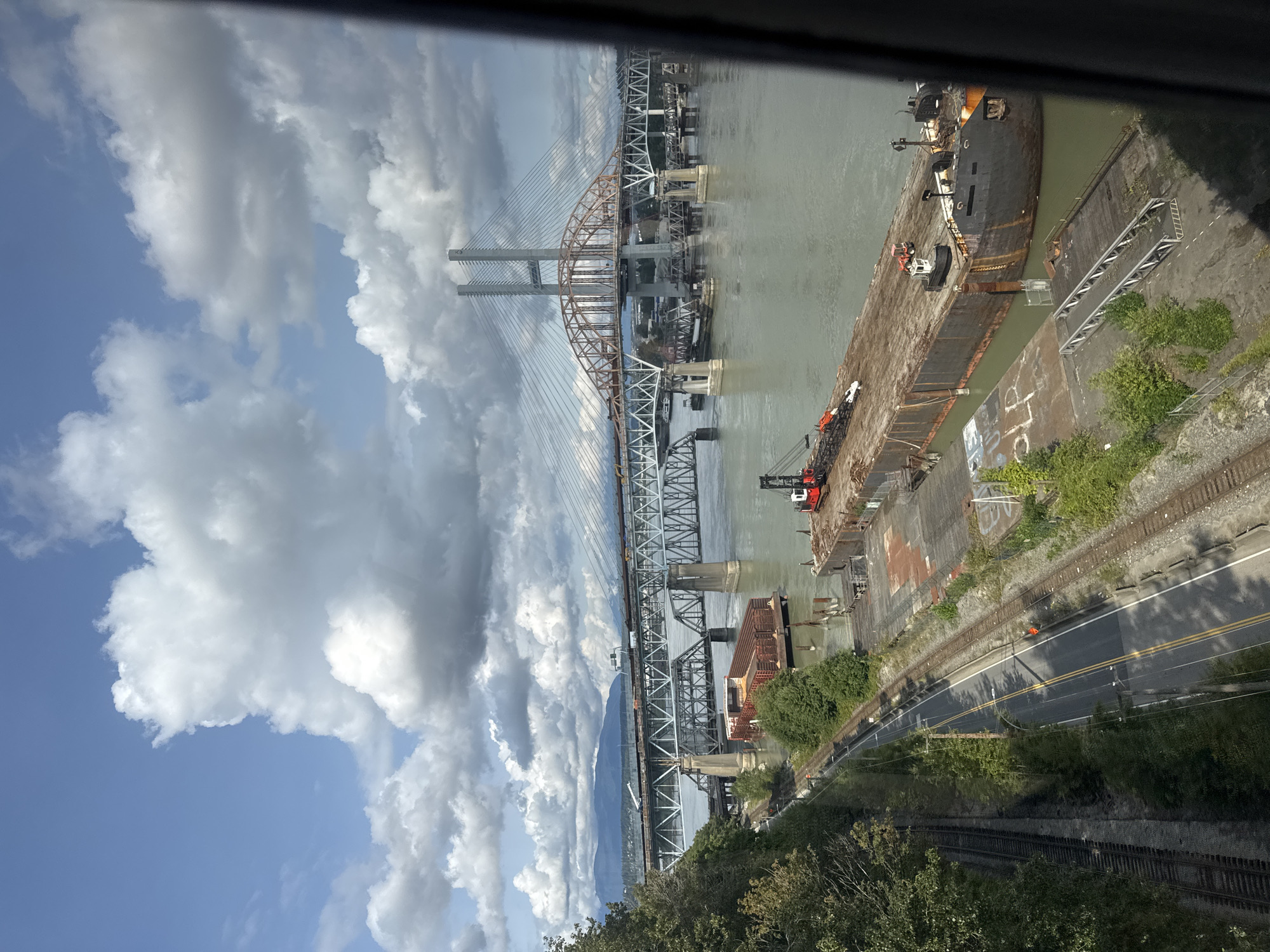 This image captures a view of the Fraser River in New Westminster, Canada, on a partly cloudy day. The foreground features a section of a large barge docked at what appears to be an industrial waterfront. The barge is dark, weathered, and appears to be carrying some cargo or debris, with a small tugboat positioned at its stern. Nearby, there are industrial structures and cranes.
A road with yellow double lines runs parallel to a set of railway tracks on the left side of the image. Adjacent to the road and tracks, there is lush green vegetation and trees.
Dominating the mid-ground and background are two distinct bridges spanning the Fraser River. The closer bridge is a large, steel truss bridge, supported by massive concrete pillars. Above and behind it, a more modern cable-stayed bridge is visible, characterized by its two tall, white towers and a distinctive orange arch structure. Trains can be seen crossing both bridges.
In the distance, a mountainous landscape is silhouetted against the sky, with the clouds casting shadows over the scene. The water of the Fraser River is a muted green-grey. The overall impression is one of a busy, industrial waterway with significant transportation infrastructure.