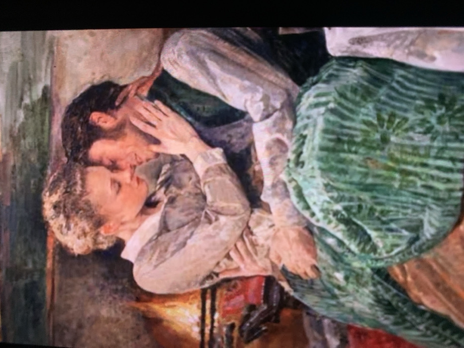 This image originates from the city of Jos, Nigeria, and depicts a detailed painting of two individuals in an intimate embrace indoors. The painting portrays a woman with light, possibly curly hair, wearing a light-colored, long-sleeved blouse, seated on the lap of a man with dark hair, dressed in a white shirt and a dark vest. Their faces are very close, almost touching, suggesting a tender moment. The woman's left hand is placed on the man's neck/shoulder, while her right arm encircles his waist. Her left leg, covered by a green patterned skirt, is draped across his lap. The man's arms are around her, with his left hand visible on her upper thigh. In the background to the left, a fireplace with visible orange and yellow flames and a dark metal grate provides warm illumination. A small red object is partially visible near the fireplace base. A white surface, possibly a tablecloth, is visible on the far right. The overall artistic style features distinct brushstrokes.