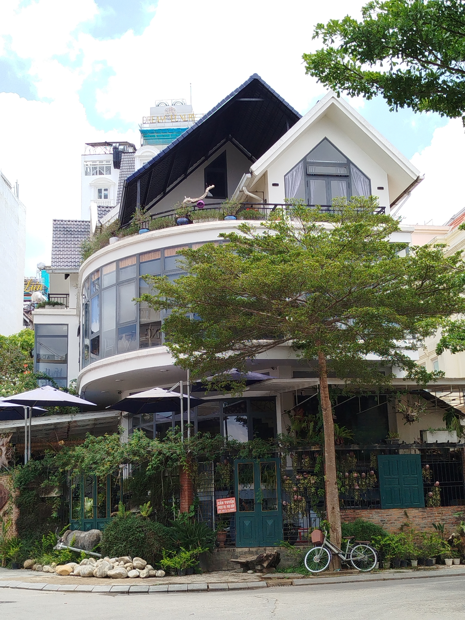 The image depicts a three-story building in Da Lat, Vietnam, characterized by a mix of architectural styles. The building’s lower level features large glass windows and a canopy with dark blue umbrellas, partially obscured by lush greenery and a bicycle leaning against a tree. A decorative display of rocks and foliage adorns the sidewalk. The middle level showcases a curved facade with reflective windows. The upper level includes a gabled section with a triangular window and a dark-roofed, open balcony area. The building is nestled amidst other structures, with the "DREAM LUXURY" sign visible atop a taller building in the background. The sky is partially cloudy. The scene is captured from a ground-level perspective, focusing on the building's architectural details and its integration with the surrounding urban environment. A sign with Vietnamese text is visible near the entrance.