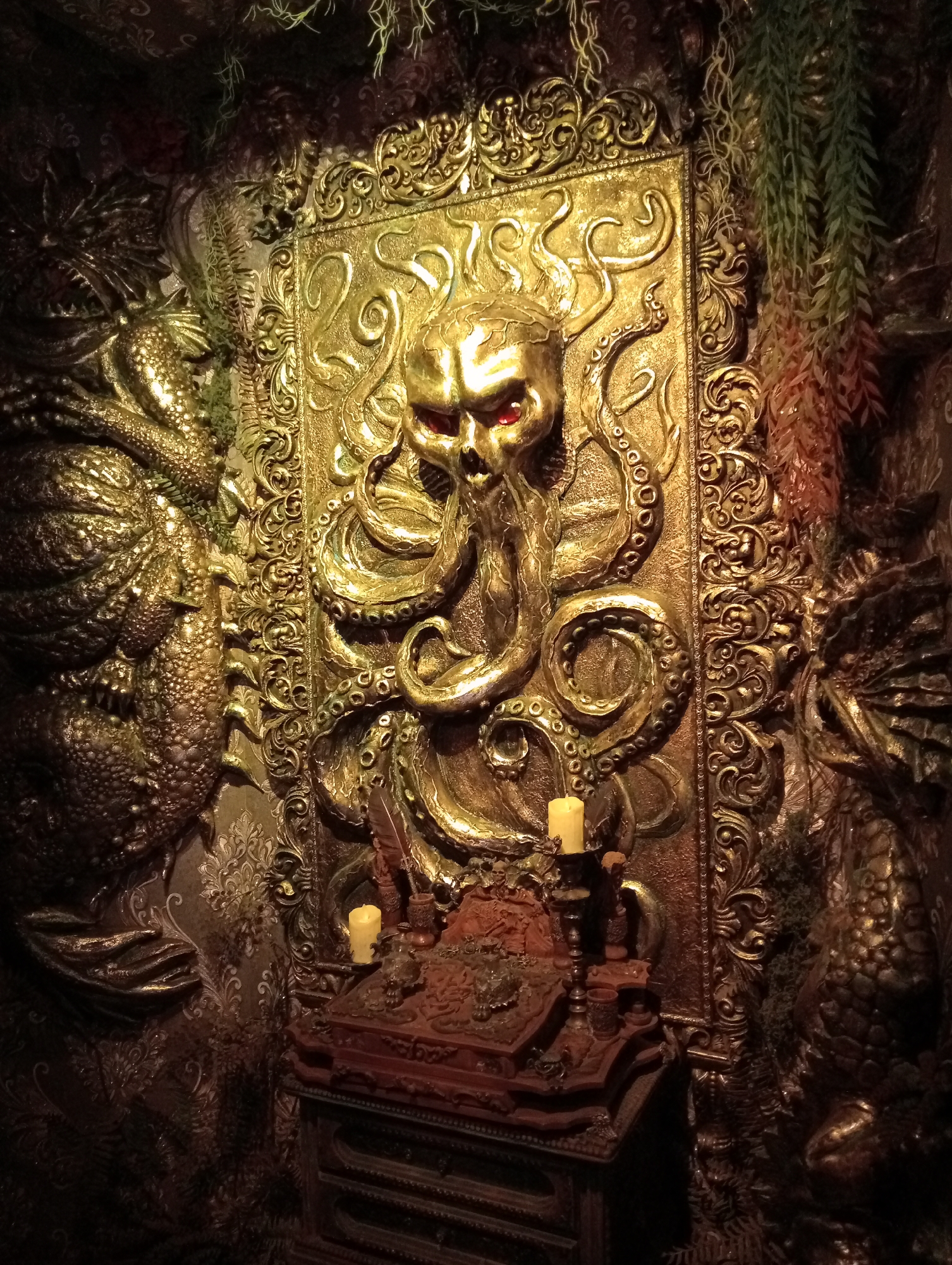 The image depicts an elaborately decorated wall display in a dimly lit, possibly gothic or steampunk-themed environment. The central focus is a large, golden, three-dimensional relief featuring a skull with red eyes, intertwined with octopus tentacles. The relief is framed by ornate golden scrollwork. Flanking the central relief are large, textured, golden dragon-like or reptilian figures. In front of the relief, on a dark wooden stand, are two lit yellow candles in ornate holders, and what appears to be a decorative book or box. Hanging plants, some appearing artificial, in shades of green and reddish-brown, are placed on either side of the central relief, adding to the atmospheric decor. The overall impression is one of dark fantasy and opulence.
There is no information in the image or its crops to suggest a location in Saint Petersburg, Russia.