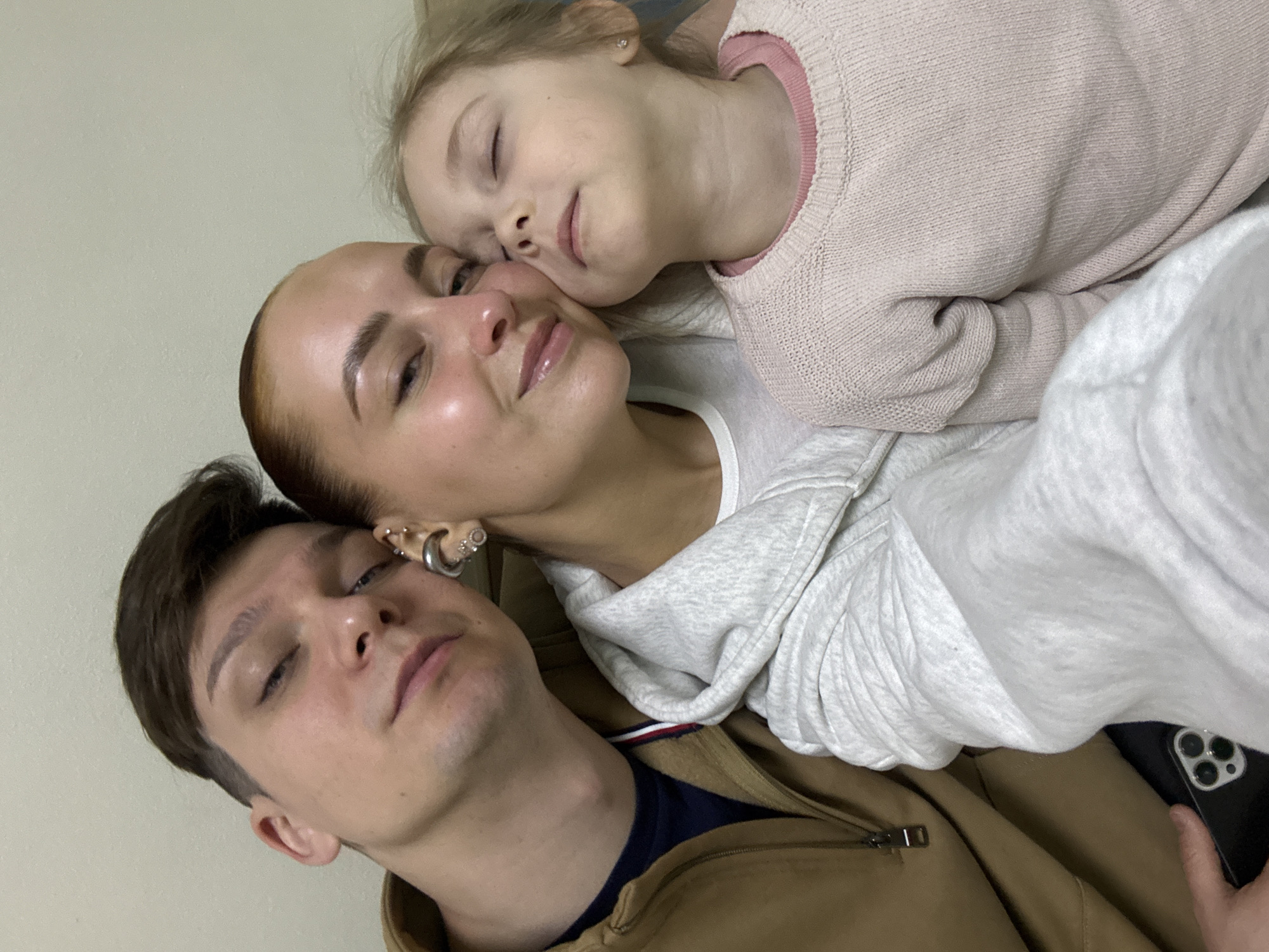 A family of three, appearing to be a man, woman, and young girl, are captured in a close-up, angled shot, resting together in an indoor setting in Chaikovsky, Russia. The man is at the bottom left of the frame, lying down with his eyes mostly closed. He has short brown hair and is wearing a brown jacket over a dark blue shirt with a red and white striped collar detail.
Next to him, the woman lies with her head resting against his ear/head. Her eyes are also mostly closed, and she has light brown hair pulled back. She is wearing a white hoodie and her ear features multiple silver-toned earrings, including a crescent moon, a hoop, and several studs. Her face shows a serene expression.
Cuddled closest to the top right, the young girl is resting her head on the woman's shoulder or chest, with her eyes closed, suggesting deep rest or sleep. She has blonde hair and is wearing a light pink knitted sweater over a pink long-sleeved top.
The overall activity depicts a moment of intimate relaxation, perhaps a nap or quiet repose, conveying feelings of peace, comfort, and strong familial affection. A plain, light-colored wall serves as a minimalist background. In the bottom right corner, a hand holds a black iPhone with a visible triple camera setup. No discernible text is visible in the image.