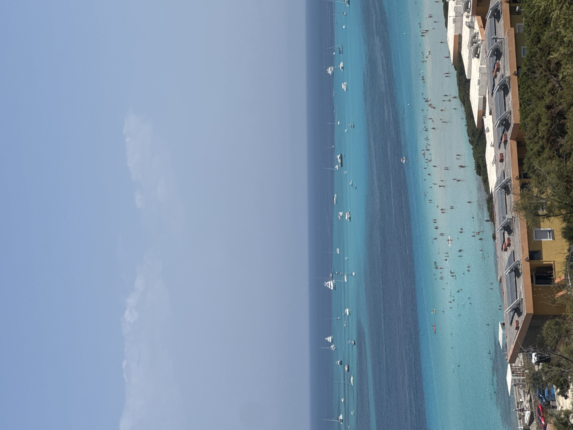 An aerial, eye-level, full shot captures a panoramic view of a vibrant seascape bordering a coastal town in Stintino, Italy. The upper portion of the frame is dominated by a clear, pale blue sky with a few wispy white clouds. Below, the deep blue of the sea transitions into a shallow, luminous turquoise closer to the shore, creating a striking contrast. Numerous sailboats and yachts are anchored or cruising in the deeper waters, their white sails and hulls dotting the expanse.
The shallow turquoise water is teeming with people enjoying the beach and swimming. Small figures are visible wading, swimming, and interacting in the water. Some are on paddleboards or small boats. Along the immediate coastline, a few structures with flat roofs are visible, adorned with solar panels. These buildings appear to be part of a low-rise resort or residential area, with some vegetation growing around them. In the foreground, a dense green bushy area is visible, suggesting a natural landscape bordering the developed area. Several cars, including a red sedan, a dark blue hatchback, and a white vehicle, are parked on a paved area near the buildings. The scene depicts a lively and picturesque coastal day in a Mediterranean setting.