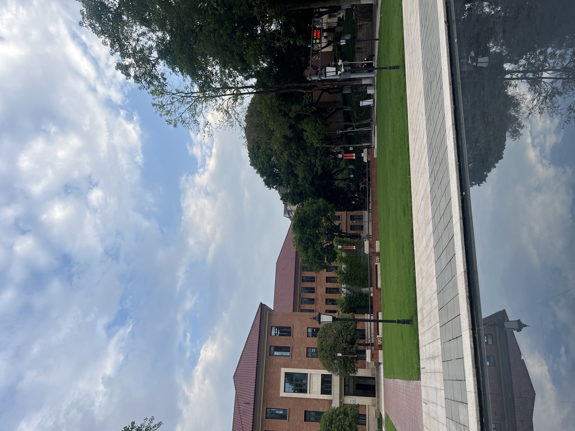 This media file presents an outdoor urban or campus scene in Yuzhong District, China, characterized by modern architecture, lush greenery, and a prominent reflective surface. The image is taken on a bright, overcast day with a blue sky partially covered by large, fluffy white clouds.
In the foreground and midground, there are several brick buildings with reddish-brown tiled roofs and numerous dark-framed windows. These buildings exhibit a mix of traditional and contemporary design, featuring large, dark glass panels on some sections. A small white sign with a black "S" is visible near an entrance on one of the buildings.
The scene is dominated by well-maintained green lawns and numerous mature trees with dense, dark green foliage, providing ample shade. Paved walkways made of light gray, dark gray, and reddish-brown square tiles run alongside the lawns and buildings.
A striking feature is a long, dark, highly reflective surface, likely a calm pool of water or a polished dark wall, which perfectly mirrors the sky, clouds, and surrounding trees and buildings. This reflection creates a sense of depth and symmetry.
In the background, nestled among trees, various street fixtures are visible. These include lamp posts, a display resembling a traffic light showing red lights and Chinese characters, and numeric indicators "20" and "60," possibly speed limits or other regulations. There's also a red circular sign, likely indicating a prohibition, and another red rectangular sign. No people or specific activities are visible, suggesting a quiet moment in the area.