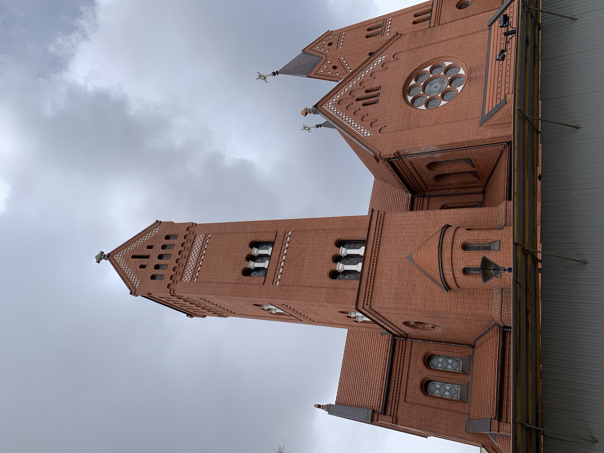 This media file captures the imposing exterior of a large brick church, identified as the Church of Saints Simon and Helena in Minsk, Belarus. The photograph is taken from a low angle, looking upwards at the architectural structure against a backdrop of a heavily overcast, cloudy sky.
The church is constructed predominantly from reddish-brown brick, exhibiting distinct Gothic Revival architectural features. A tall bell tower dominates the left side of the image, culminating in a pointed spire topped with a cross. Other spires or turrets, visible towards the upper right, also feature ornate finials, possibly made of patinated metal, with a greenish hue.
Notable architectural details include intricate brickwork patterns, decorative white criss-cross bands adorning the gables and cornices, and various window designs. These include tall, arched windows with white, column-like mullions, smaller rectangular windows, and a large circular rose window with multiple glazed panes prominently featured on a side facade. The roofs are tiled, with some sections appearing dark gray and others complementing the brickwork. On the right edge of the frame, a dark gray roof structure, possibly of an adjacent building or an extension, with visible support beams, can be seen.
The weather is distinctly overcast, characterized by thick gray clouds, indicating dull daylight conditions. There are no people visible, and no specific activities are taking place. No discernible text is present in the image. The focus is entirely on the historical and architectural grandeur of the church.