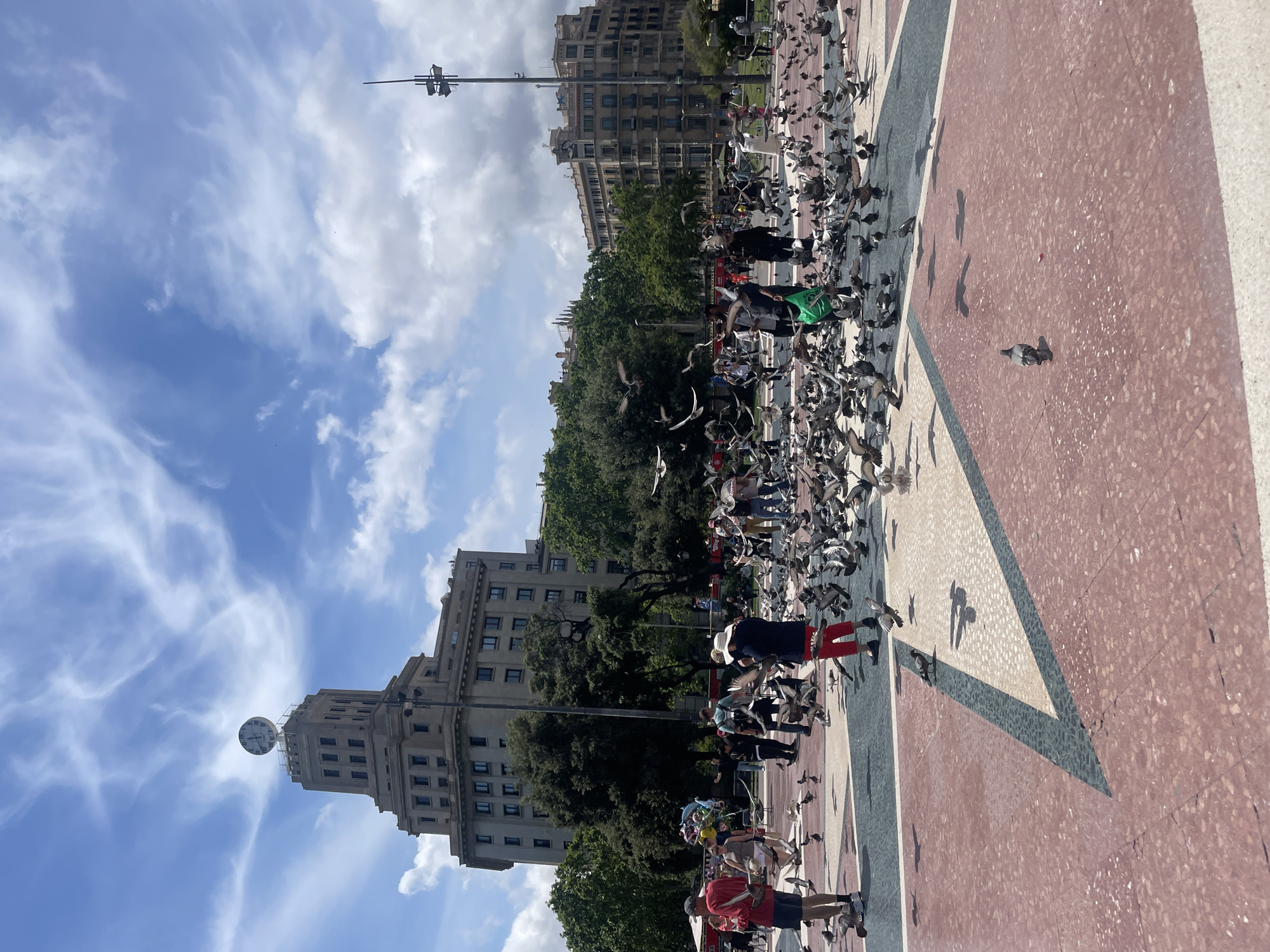 The image depicts a bustling scene in Plaça de Catalunya in Barcelona, Spain. The plaza is filled with pigeons, many of which are in flight. Several people are present, some seemingly interacting with the birds, possibly feeding them. The ground of the plaza has a patterned design in red, white, and green. Buildings, including a prominent tower with a clock, are visible in the background. The sky is blue with scattered clouds.