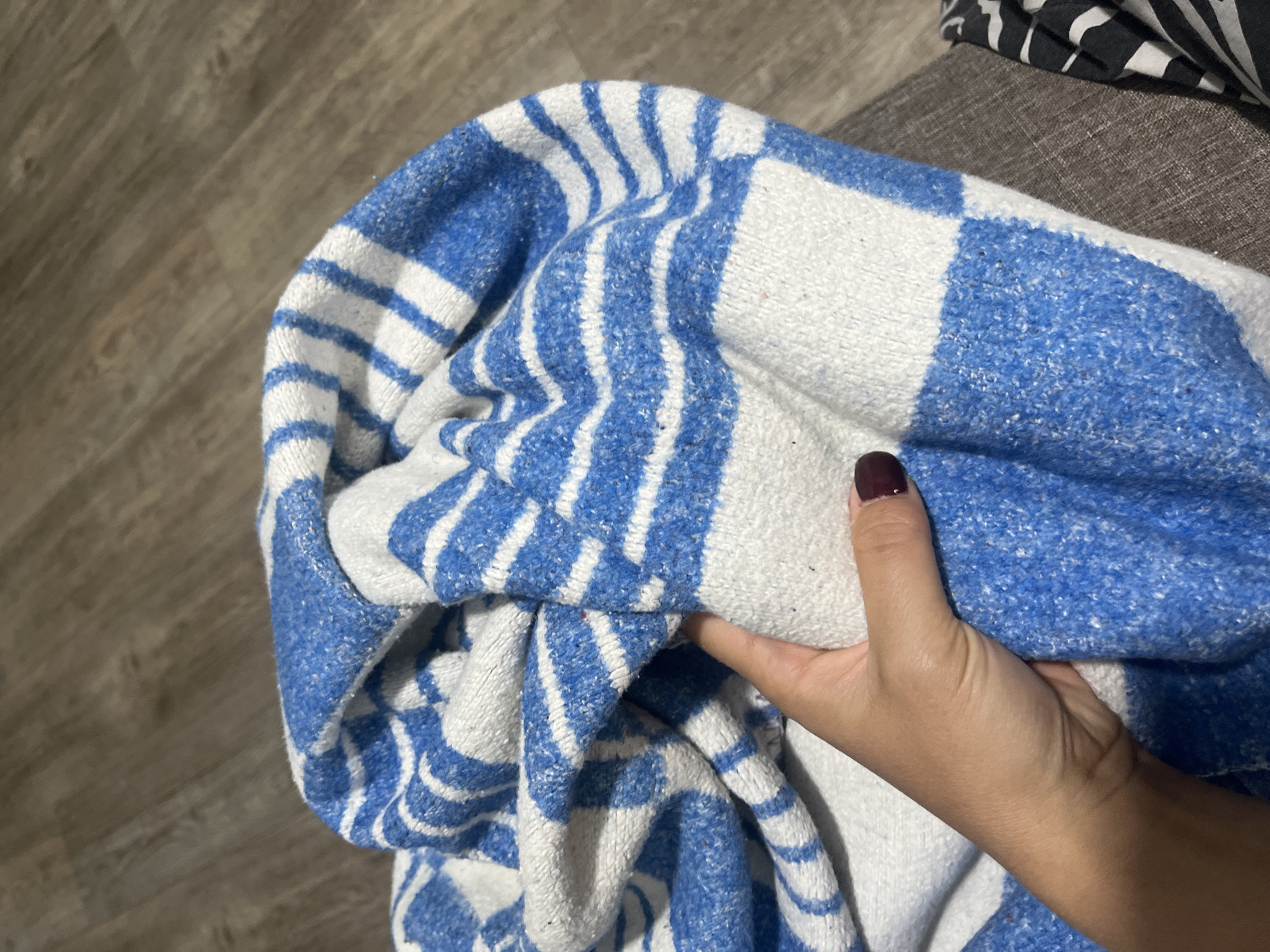 The image shows a hand holding a blue and white striped blanket. The blanket appears to be made of a soft, knitted material and is folded over itself. The hand is lightly gripping the blanket, with fingernails painted a dark color. In the background, a wooden floor is visible, suggesting an indoor setting. The scene is likely located in a residential area within the Сеніцкі сельскі Савет, Belarus, though no specific landmarks or city indicators are present in the image itself to pinpoint an exact location.