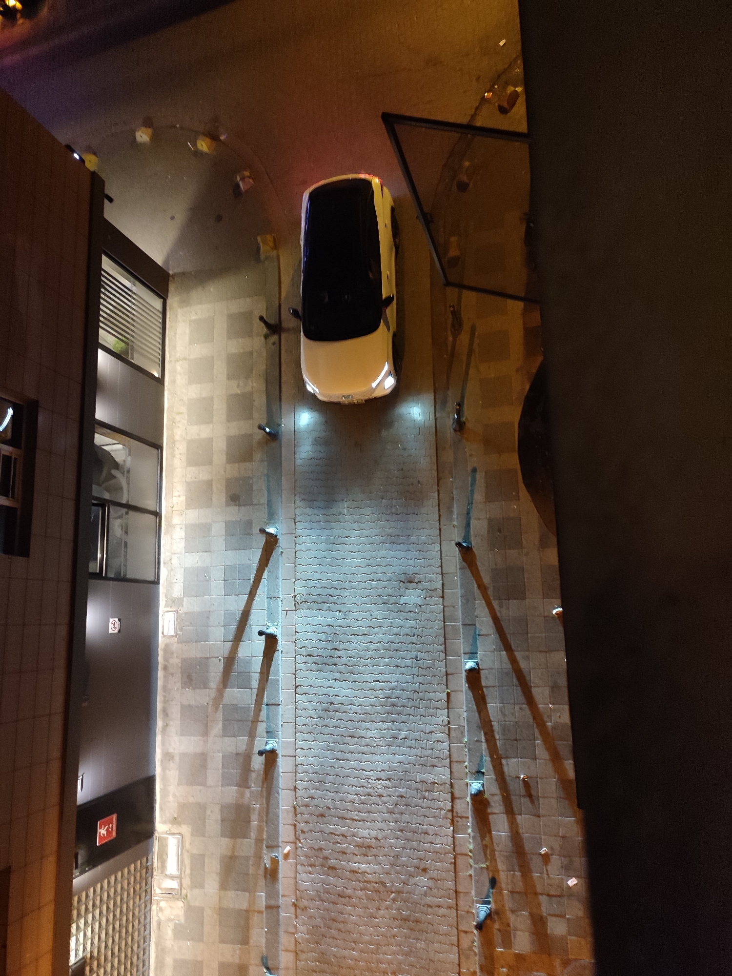 An aerial, night-time, medium-shot captures a white electric car with a black roof parked outdoors. The car is oriented with its front facing upwards and slightly to the right. The car is illuminated by its own headlights, which cast bright beams onto the ground.
The car is situated on a paved area, possibly a street or alleyway, adjacent to a building on the left. The building has a facade of brown tiles and features large windows with horizontal blinds. A dark-colored structure or wall occupies the right side of the image, obscuring a portion of the background.
The paved ground is a mix of square tiles and a textured pattern. Several dark posts are visible along the edge of the paved area, casting long shadows. The scene appears to be in an urban environment.
The image is taken from a high vantage point, looking down at the car and its surroundings. The lighting suggests artificial illumination from streetlights and the car's headlights, creating a contrast between illuminated and shadowed areas. There are no people visible in the image. The overall atmosphere is quiet and still.
The provided crops show details of the building facade with signage, the car's headlights and roof, and the textured paving stones.
The location is the street behind the Şeker Portakalı building on Caferaga Mah. Sakız Gülü Sokak in Kadıköy, Istanbul, Turkey.