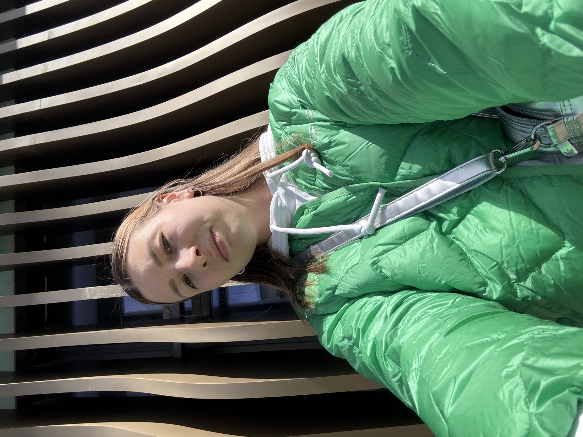 A young individual, likely female, is featured in a close-up portrait, appearing to be taking a selfie or being photographed. They have fair skin, light brown hair, and a slight, pleasant smile, looking directly into the camera.
The person is wearing a bright, shiny green puffer jacket over a white hooded garment, possibly a hoodie, visible around the neck and hood area. A silver-colored strap, belonging to a bag (likely a shoulder or crossbody bag), is visible across their chest, with a metal clasp that features a small Adidas trefoil logo. On the green jacket, near the hood, text that reads "JUST THES" is discernible.
The background consists of a repeating pattern of vertical, slightly curved slats, which appear to be part of a modern building's facade or a decorative architectural element. The lighting suggests it is bright daylight, indicating sunny weather. The overall setting implies an outdoor, urban environment. Given the provided context, this scene is set in Moscow, Russia.