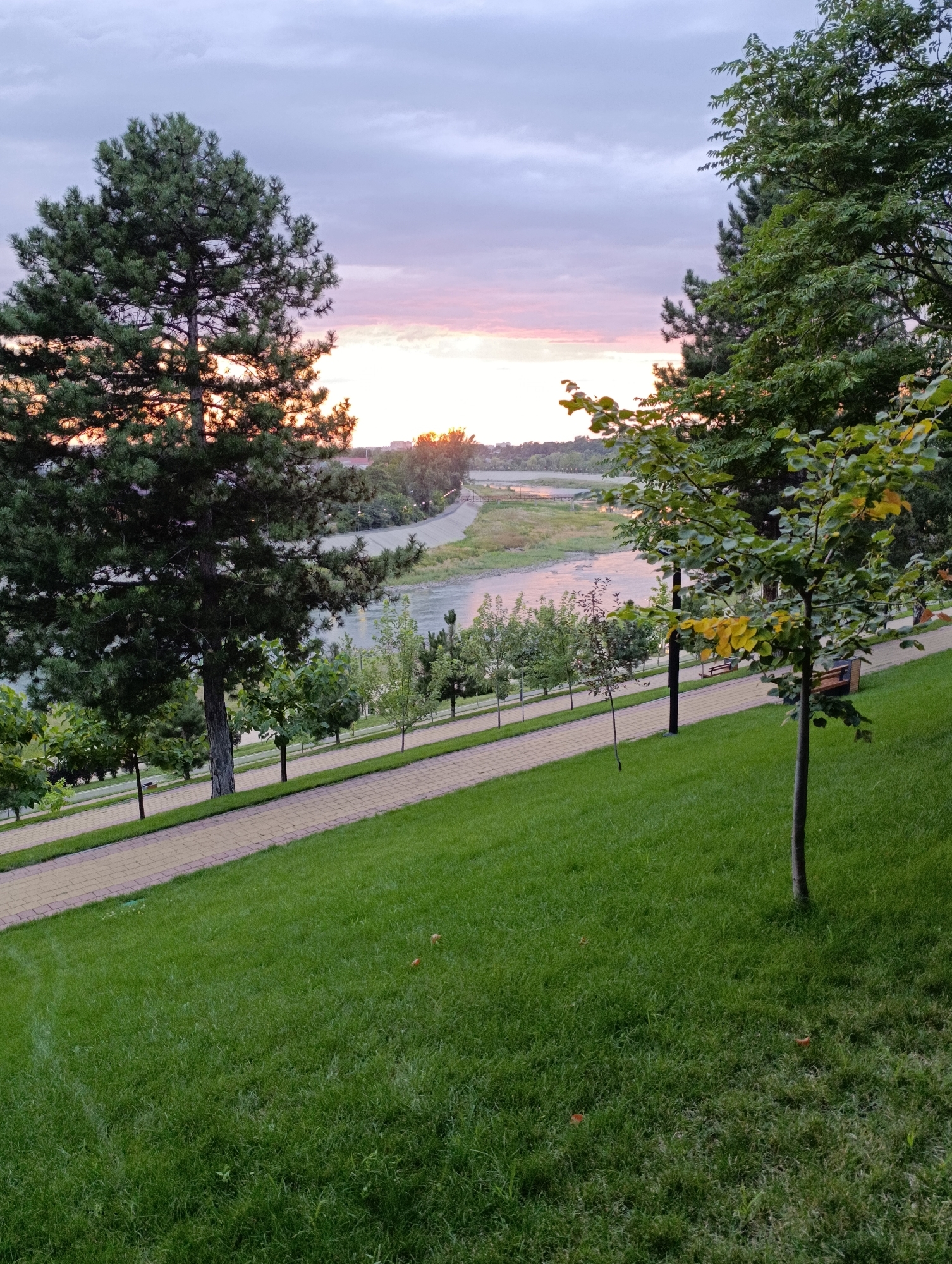 The image shows a park area with a paved walkway, a grassy slope, and trees. In the background, a body of water, likely a river, is visible with a bridge and some buildings on the far bank. The sky is overcast with hints of pink and orange suggesting a sunset or sunrise. The park appears to be well-maintained with mowed grass and ornamental trees lining the walkway. There are no people or animals visible in the scene. The overall impression is a peaceful, natural environment within a town or city. The location is specified as Kelermesskaya, Russia.